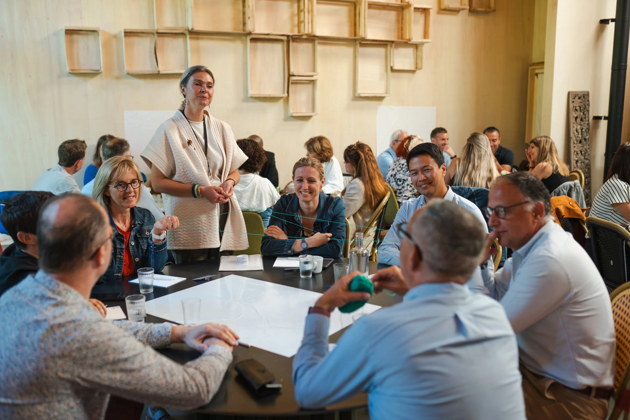 Une femme se tient debout à une table ronde entourée de personnes lors d'une réunion ou d'un atelier, dans une salle décorée avec des étagères en bois sur le mur.