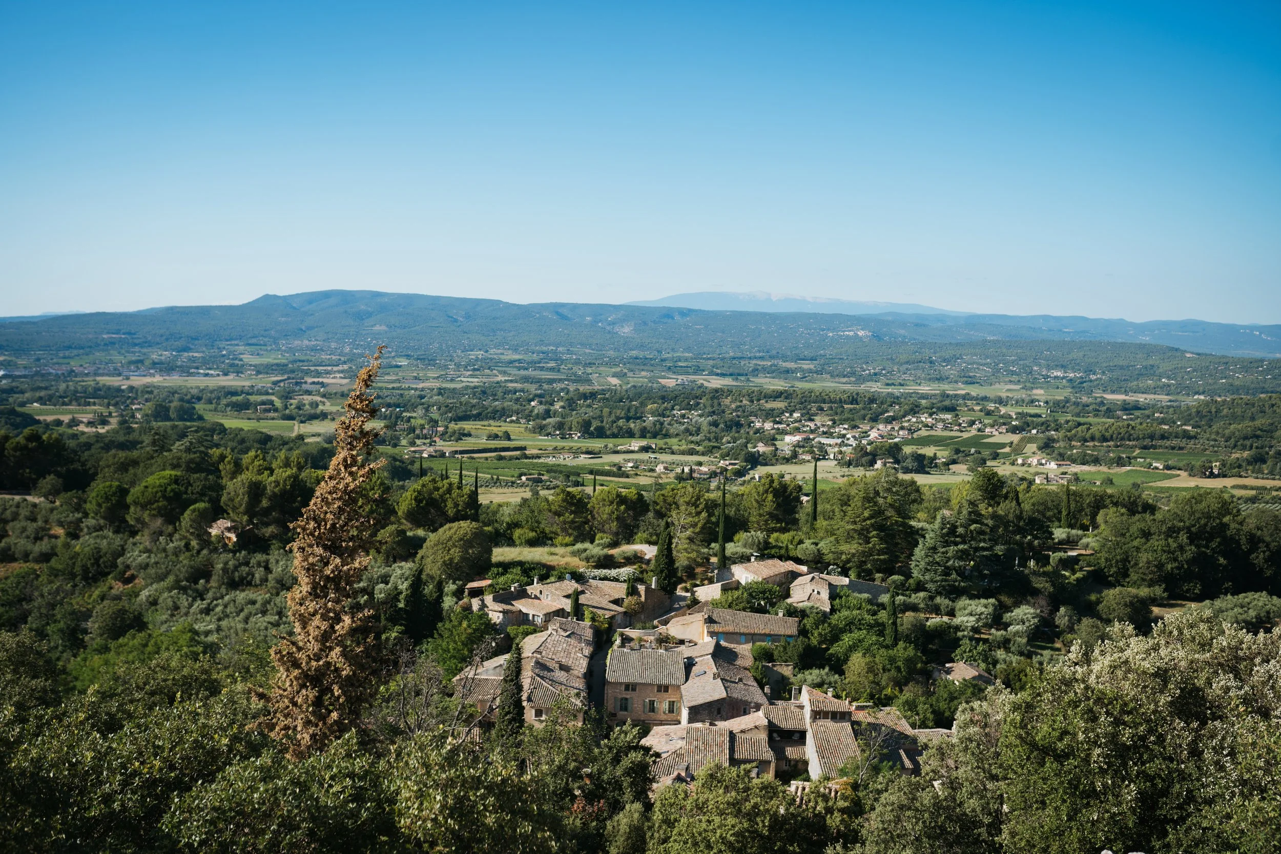 Paysage rural avec des maisons en pierre entourées d'arbres, champs fertiles et montagnes en arrière-plan sous un ciel bleu clair.