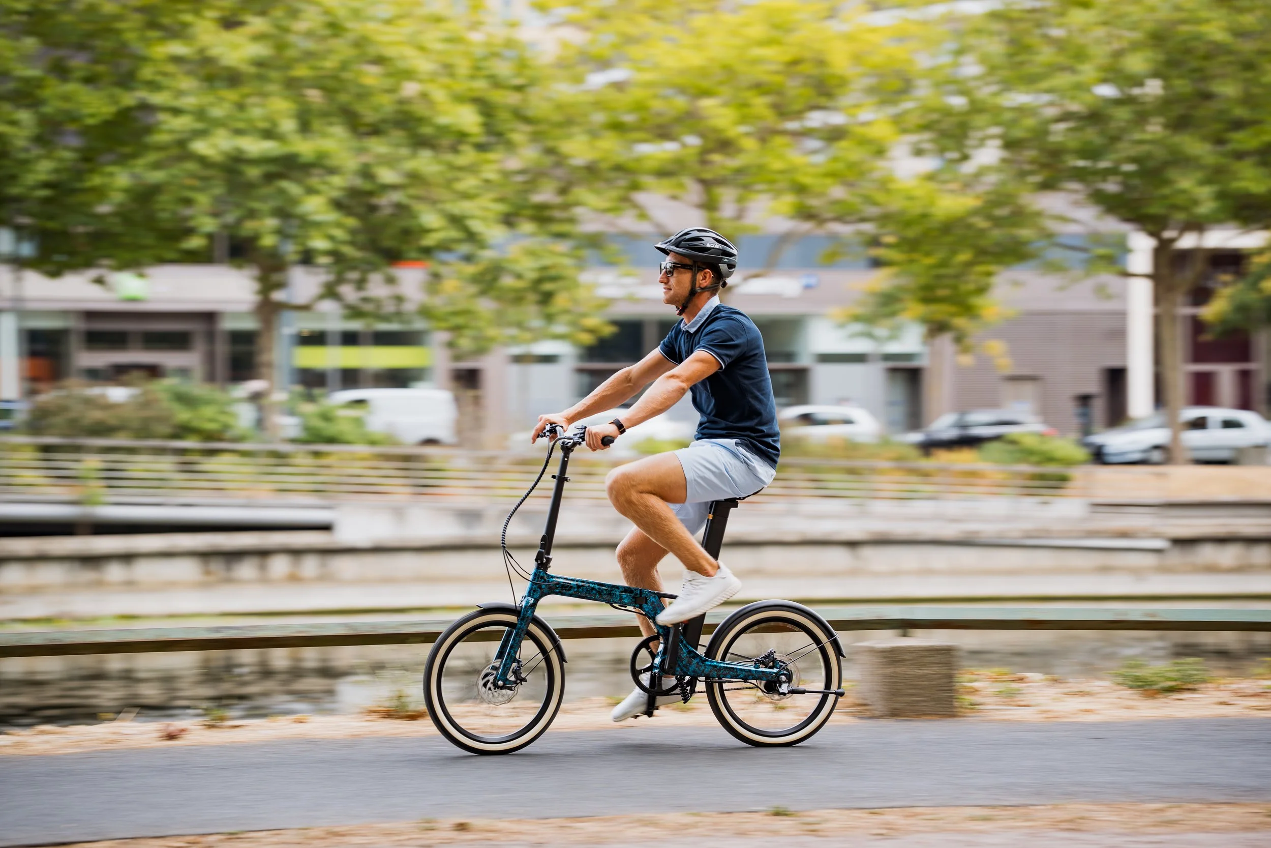 Un homme jeune portant un casque et des lunettes, pédalant sur un vélo dans une ville avec des arbres verts et des bâtiments en arrière-plan.