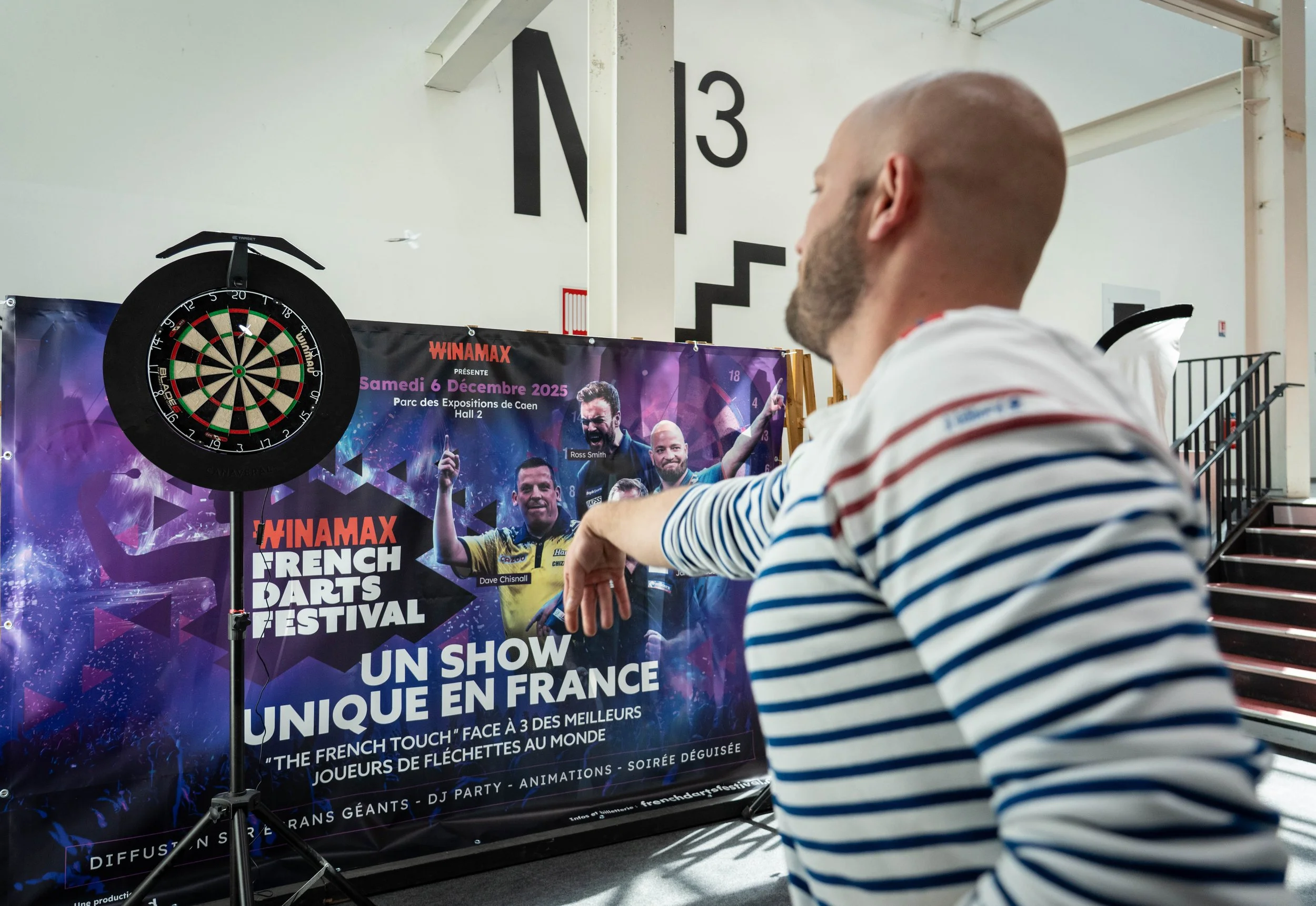 Un homme avec une tête rasée et portant un t-shirt à rayures regarde un panneau d'un festival de fléchettes françaises. À gauche, un mat de fléchettes et une bannière publicitaire sont visibles, annonçant un festival de fléchettes françaises le samedi 6 décembre 2025.