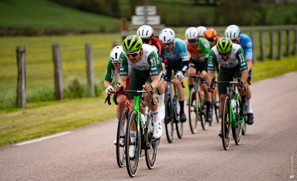 Groupe de cyclistes en course sur une route rurale avec un paysage vert en arrière-plan, portant des casques et des maillots de différentes couleurs.