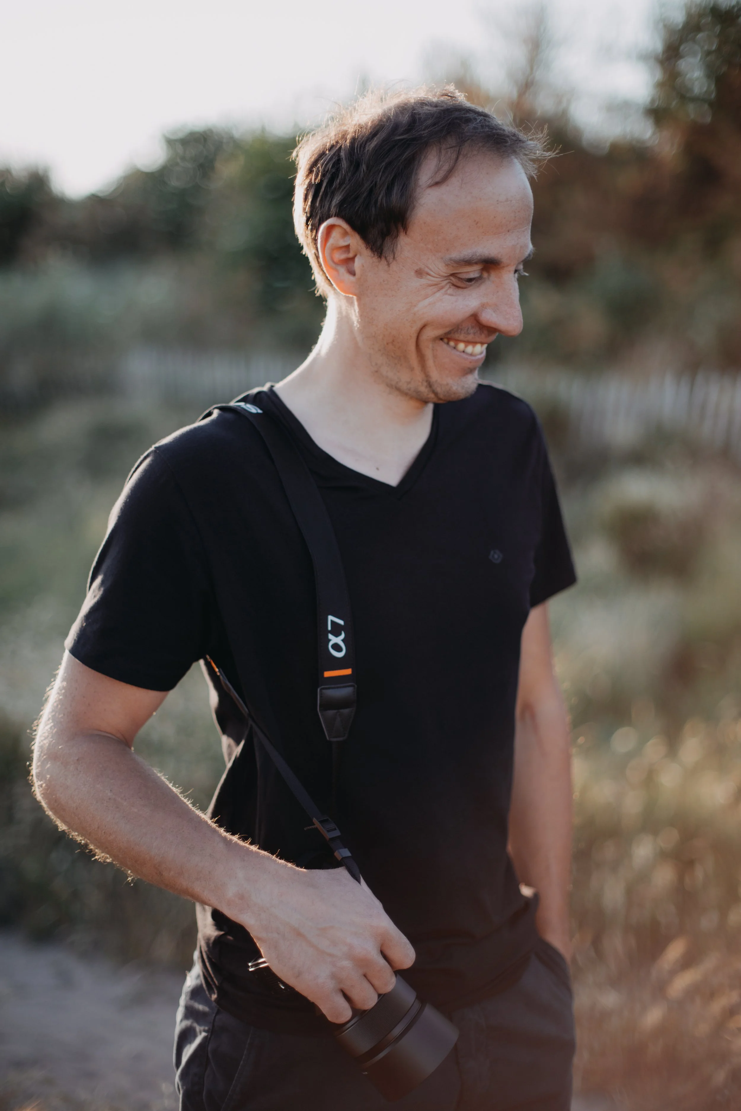 Un homme souriant en tenues noires, portant un sac à bandoulière et une bouteille d'eau, lors d'une marche en plein air au coucher du soleil.