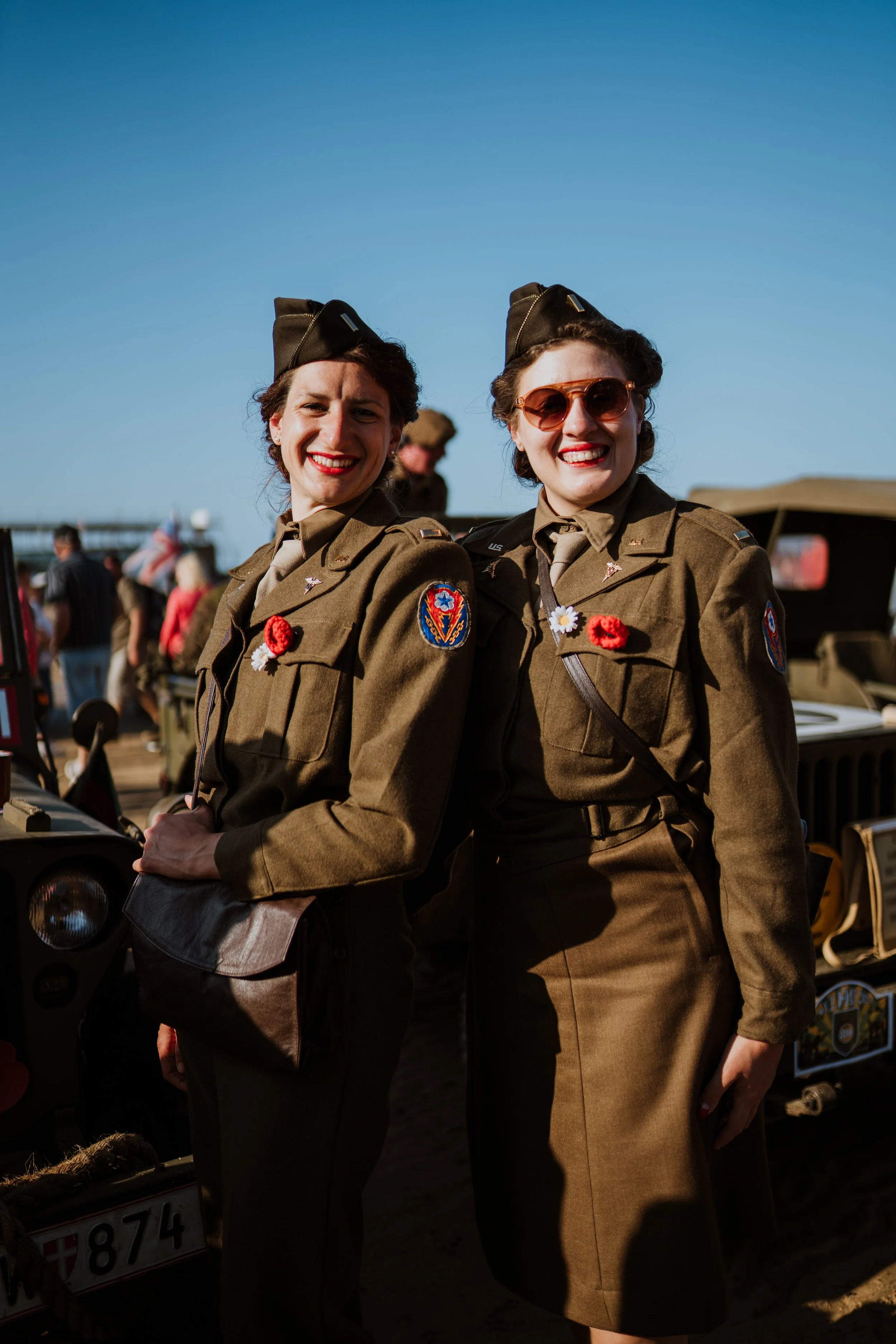 Deux femmes en uniforme militaire vintage posant en plein air, souriant, avec des sourires éclatants, portant des badges et des accessoires de mode rétro, sous un ciel clair et ensoleillé.