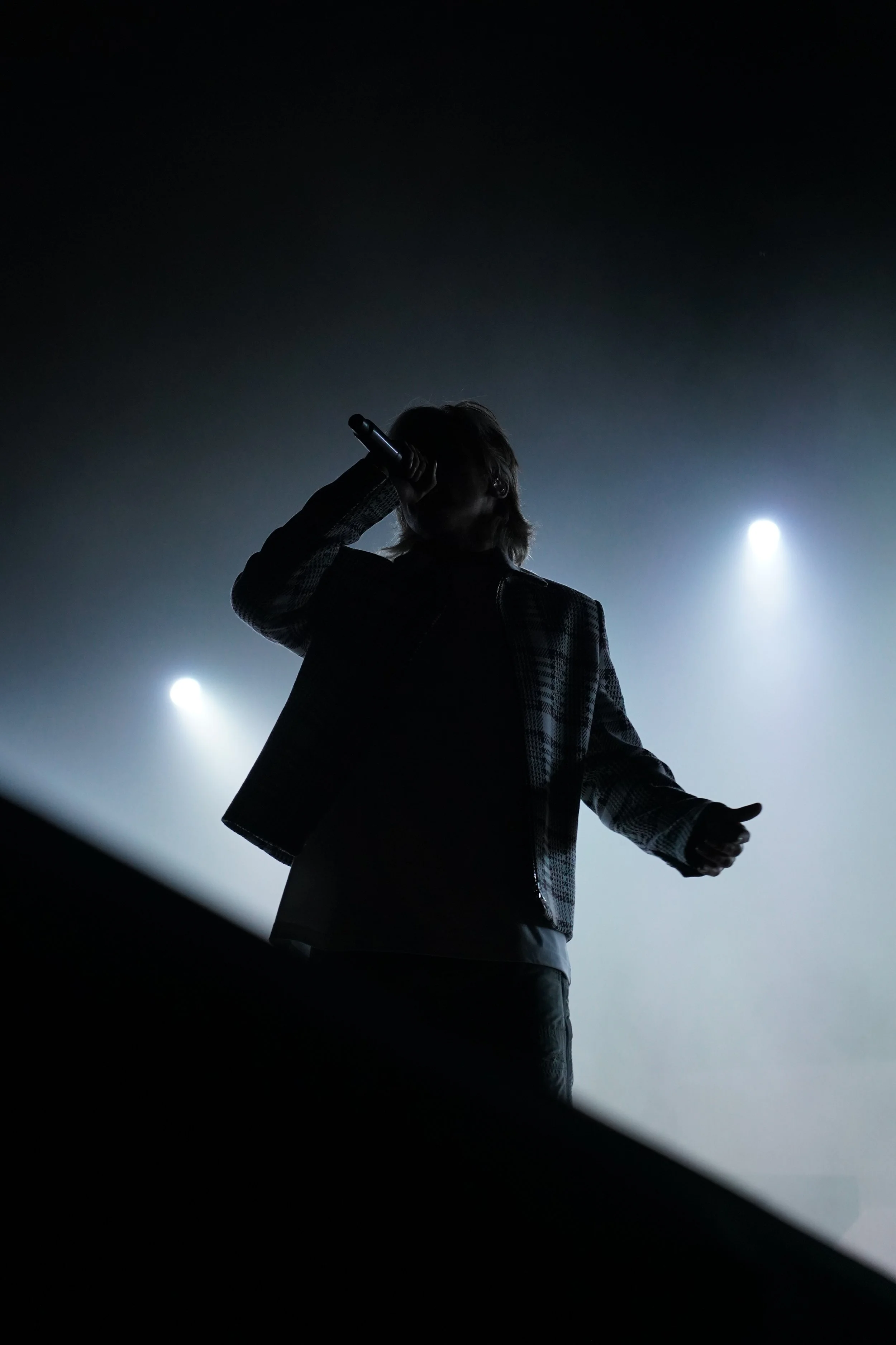 Une silhouette d'une personne chantant ou parlant, tenant un microphone, avec un arrière-plan sombre et une lumière vive derrière.