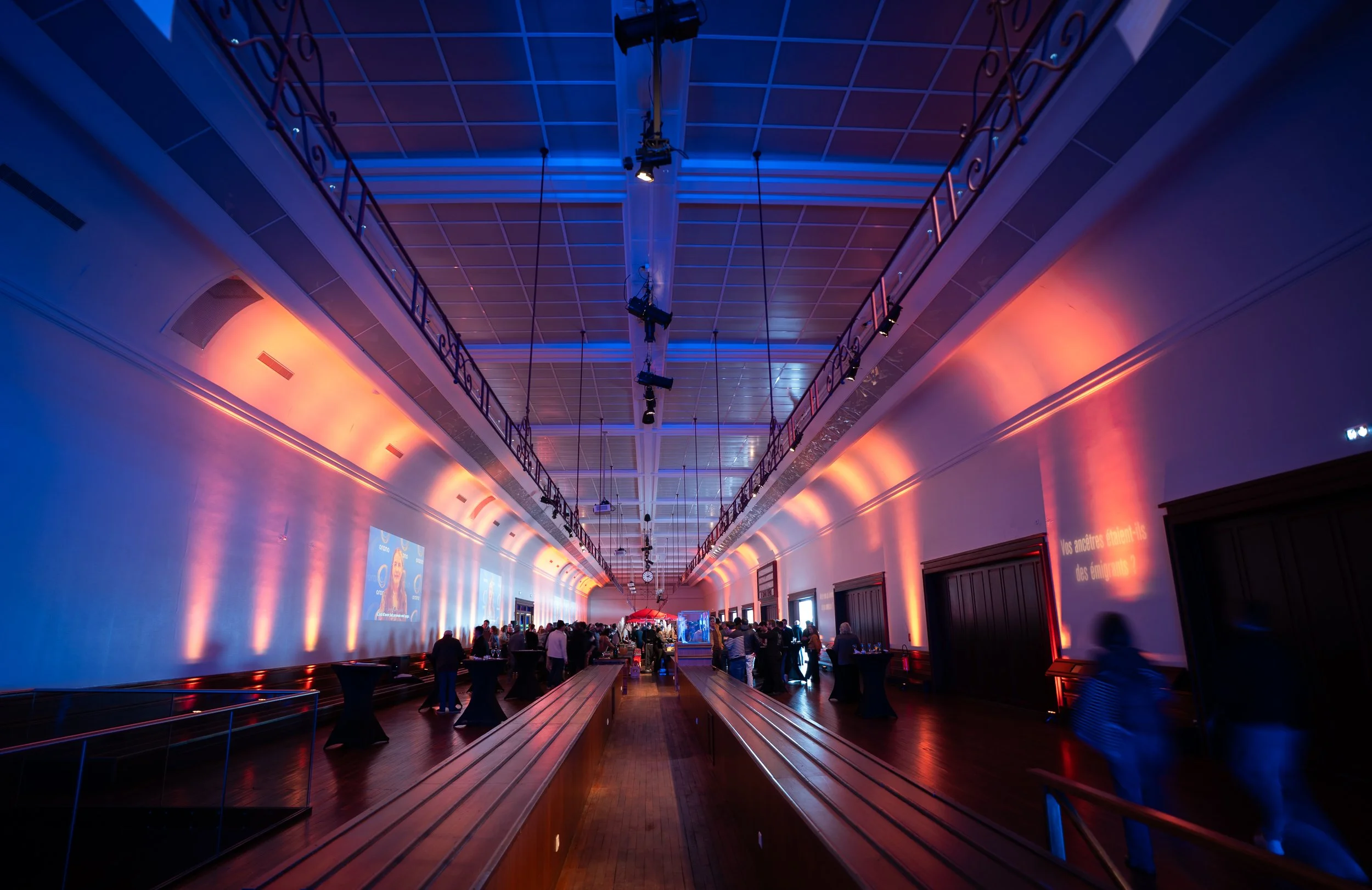 Une grande salle avec un plafond équipé d'éclairage bleu et orange, remplie de personnes assises et debout, avec des écrans et des projecteurs suspendus.