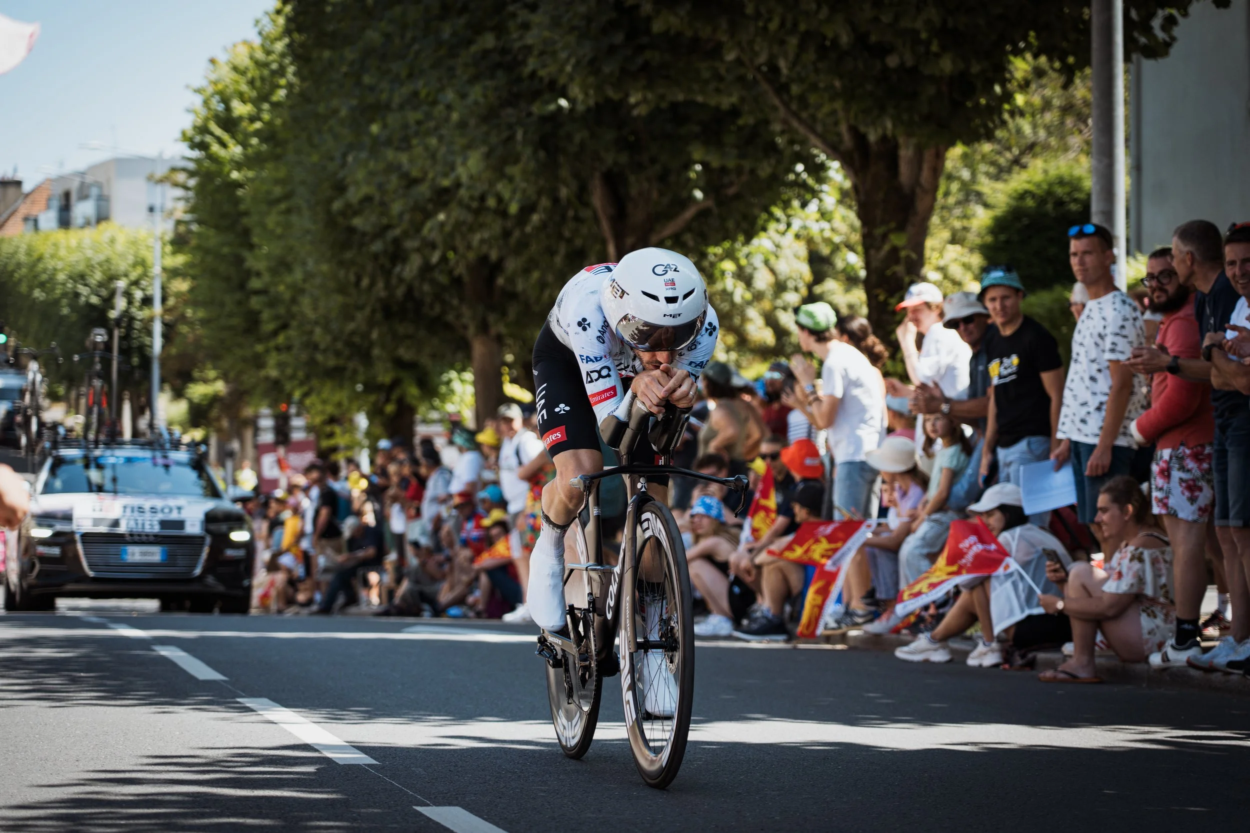 Un cycliste en tenue de course, portant un casque blanc, avançant à vélo lors d'une compétition, avec une foule de spectateurs assis et debout qui l'encouragent, certains tenant des drapeaux, sous un arbre ombragé, en milieu urbain.