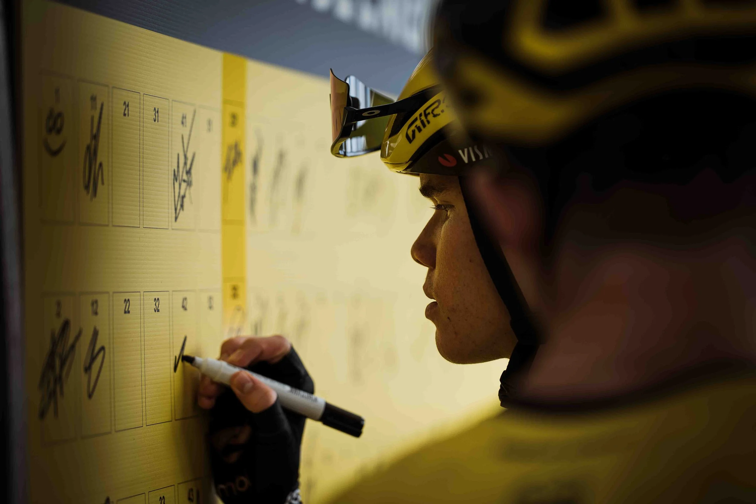 Un homme portant un casque de cyclisme jaune écrit avec un marqueur noir sur un tableau jaune.