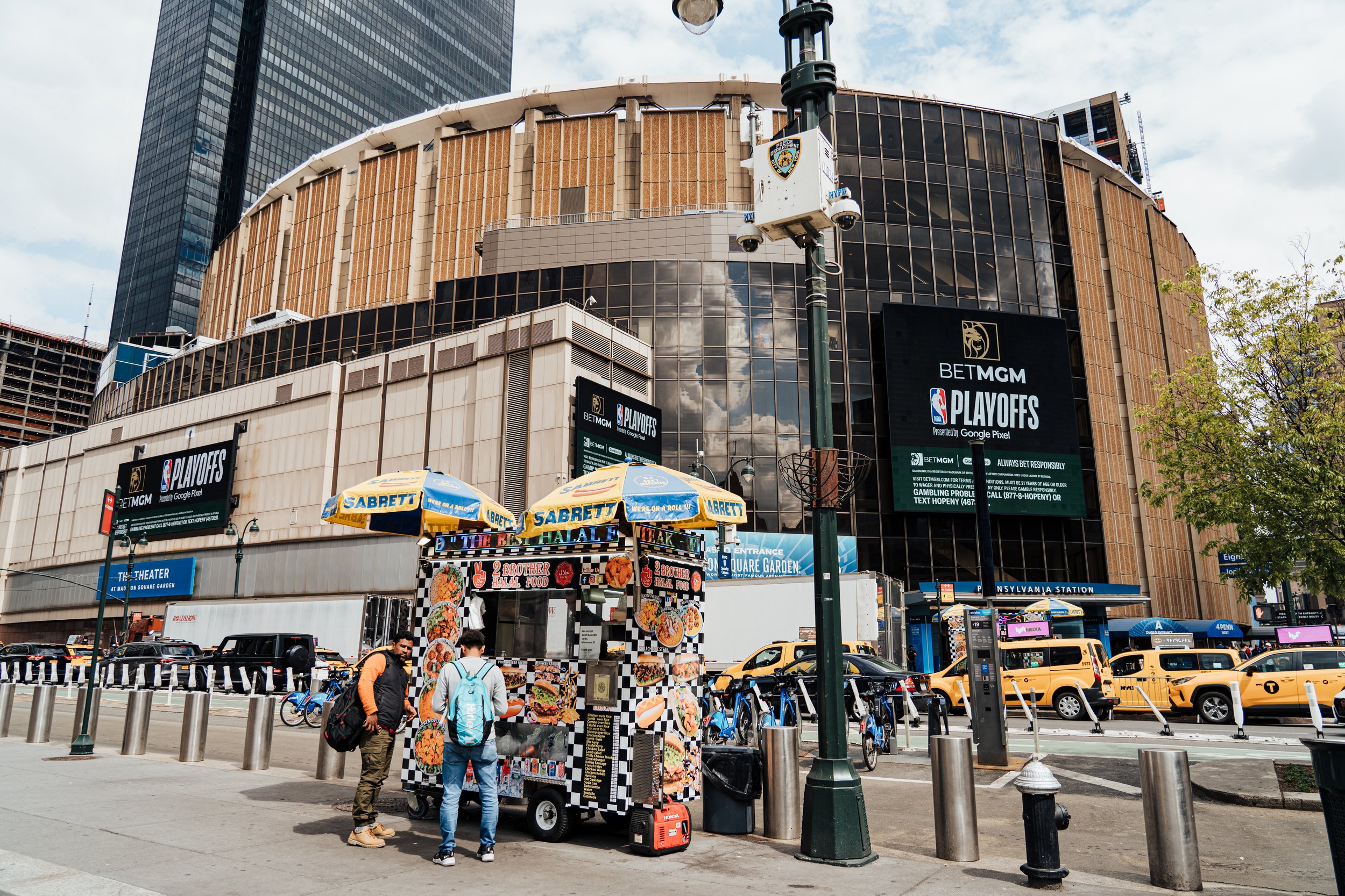 Vue d'une zone urbaine avec un bâtiment en verre et en pierre, des panneaux d'affichage pour les séries NBA Playoffs de BETMGM, une station de métro NY Penn, des stations de vélos, des taxis jaunes et une tente de vente de nourriture avec des gyro de fruits de mer.