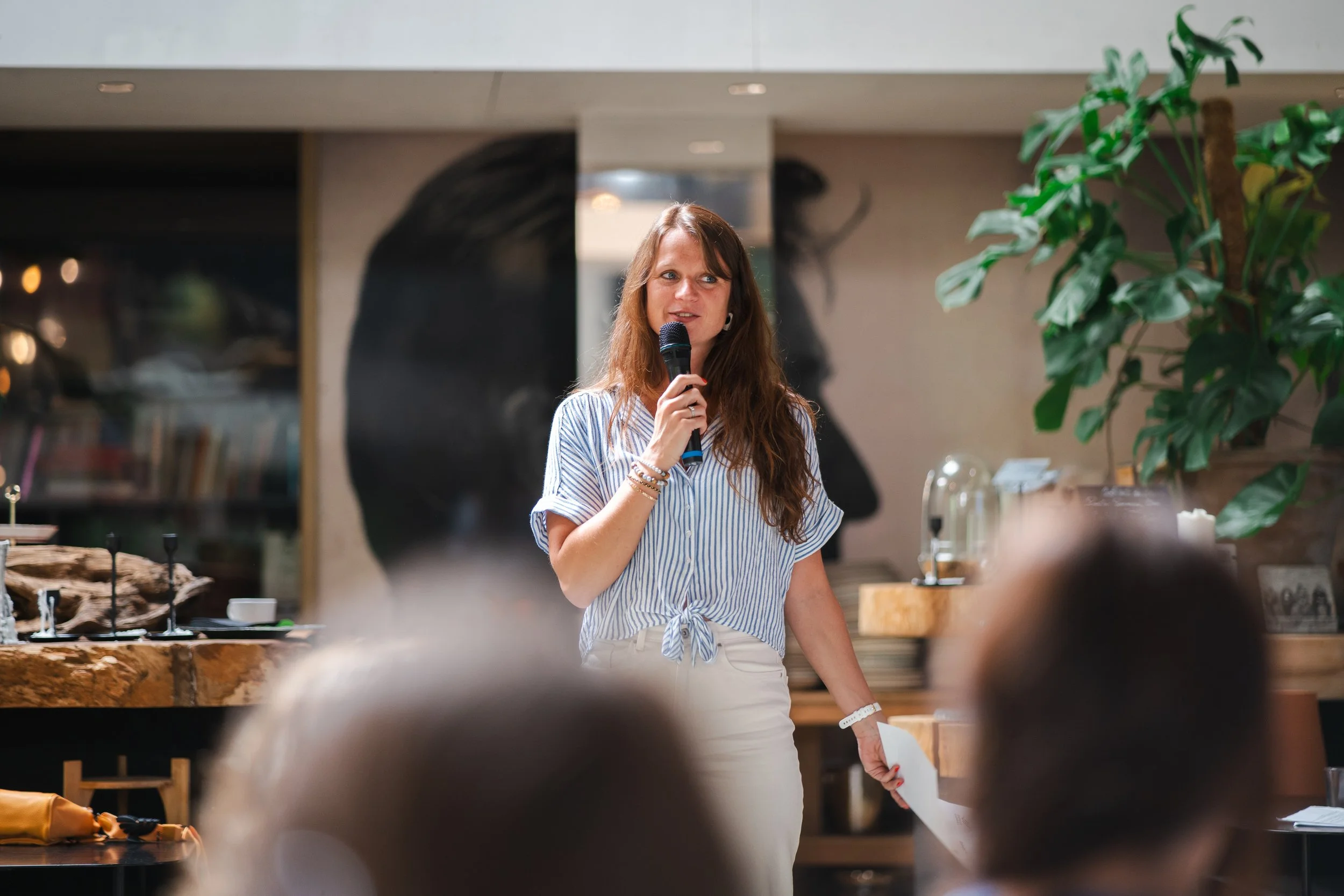 Une femme avec des cheveux bruns longs, portant une chemise rayée bleue et blanche, tenant un micro, parlant dans un environnement intérieur avec décor vert et cadre artistique.