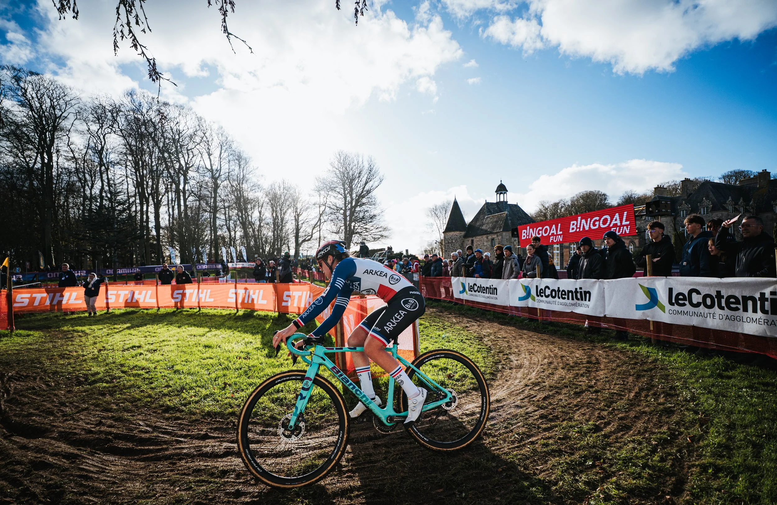 Un cycliste en compétition lors d'une course dans un parc avec arbres et un bâtiment historique en arrière-plan, sous un ciel partiellement nuageux.