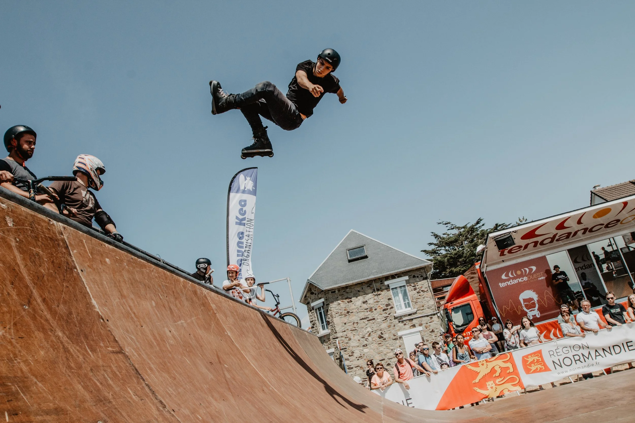 Skateur effectuant un saut dans une compétition de skate en extérieur, avec un public et des caméras en arrière-plan, sous un ciel dégagé.