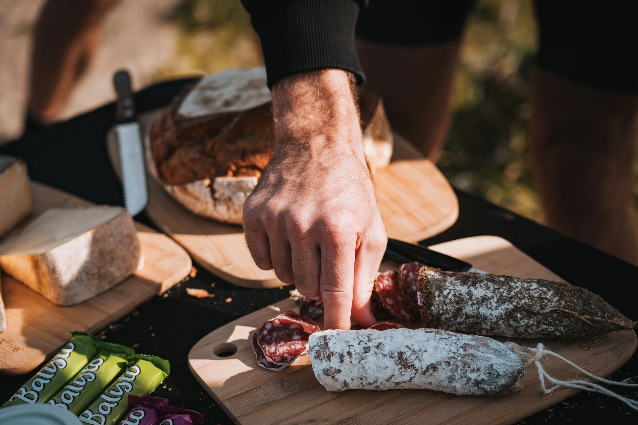 Une main manipule des morceaux de charcuterie sur une planche en bois, entourée de différentes charcuteries et snacks.