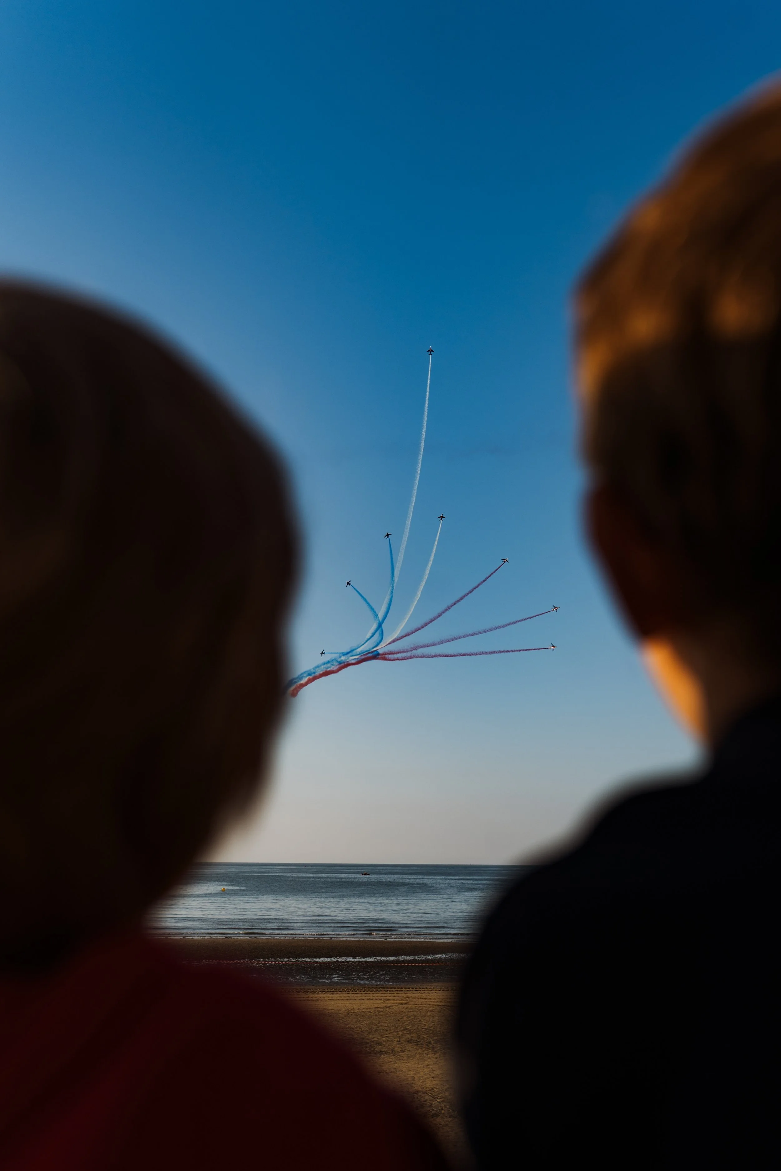 Un groupe de personnes regarde un ciel clair où plusieurs avions laissent des traces colorées, en contrebas d'une plage et de la mer, lors d'une arrivée ou d'un spectacle aérien.