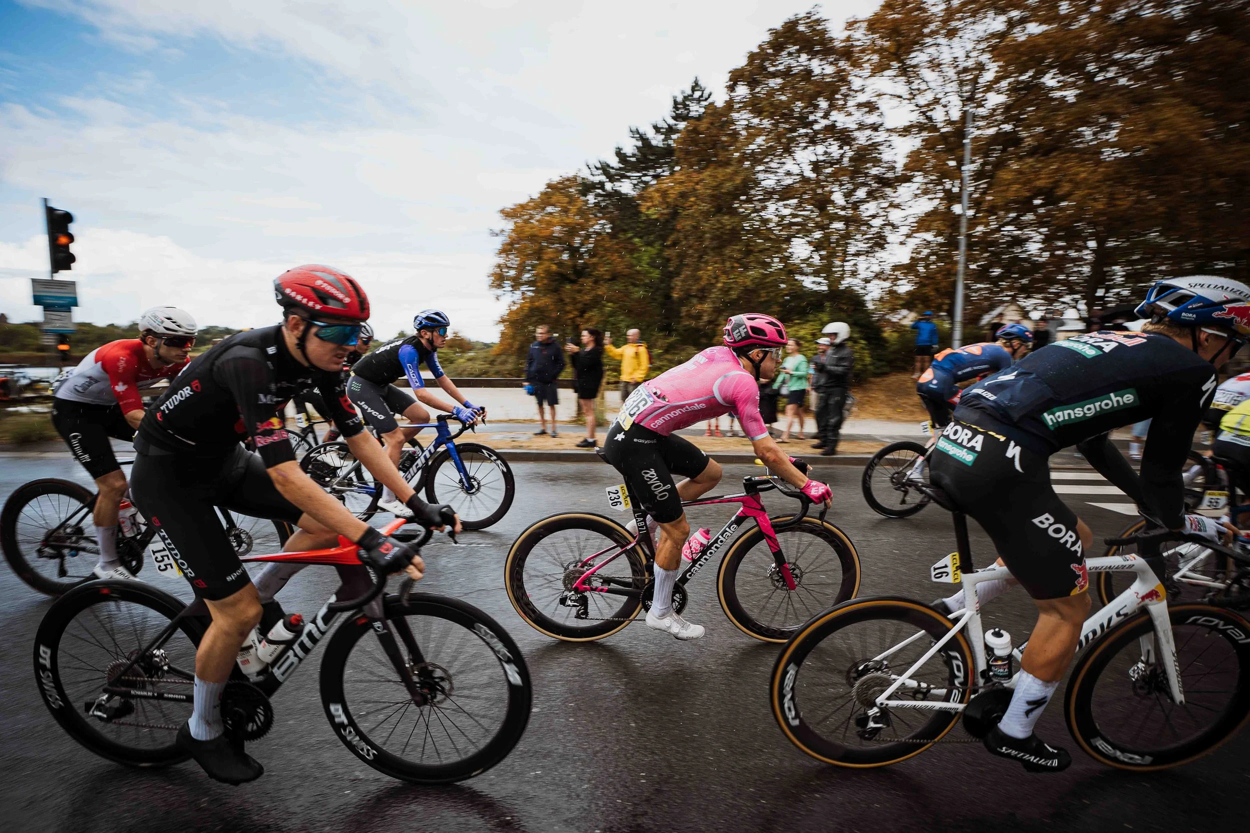 Groupe de cyclistes participant à une course sur une route humide par temps gris, avec des arbres aux couleurs automnales et quelques spectateurs en arrière-plan.