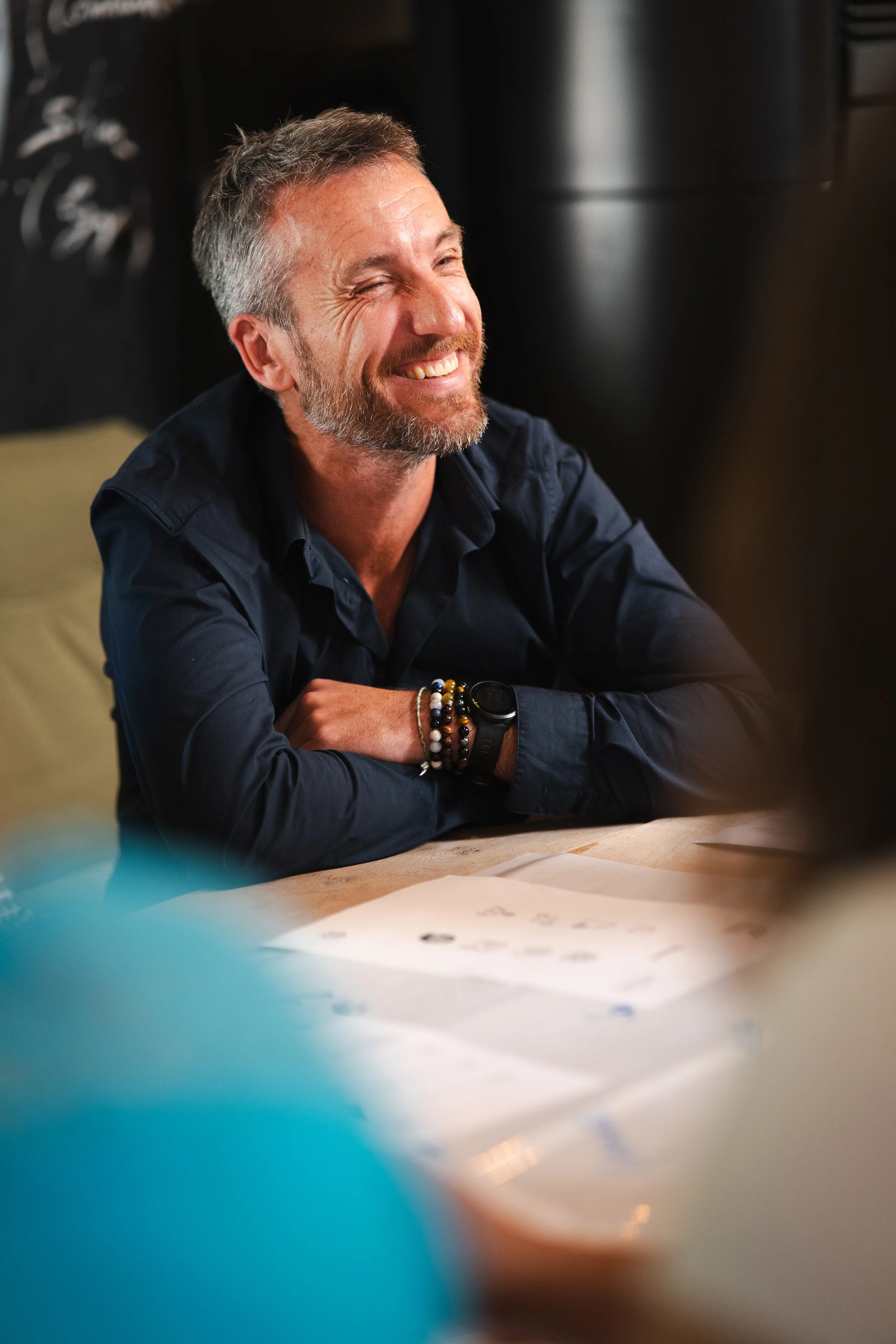 Un homme souriant, assis à une table, portant un bracelet et une montre, avec des papiers devant lui dans un environnement intérieur