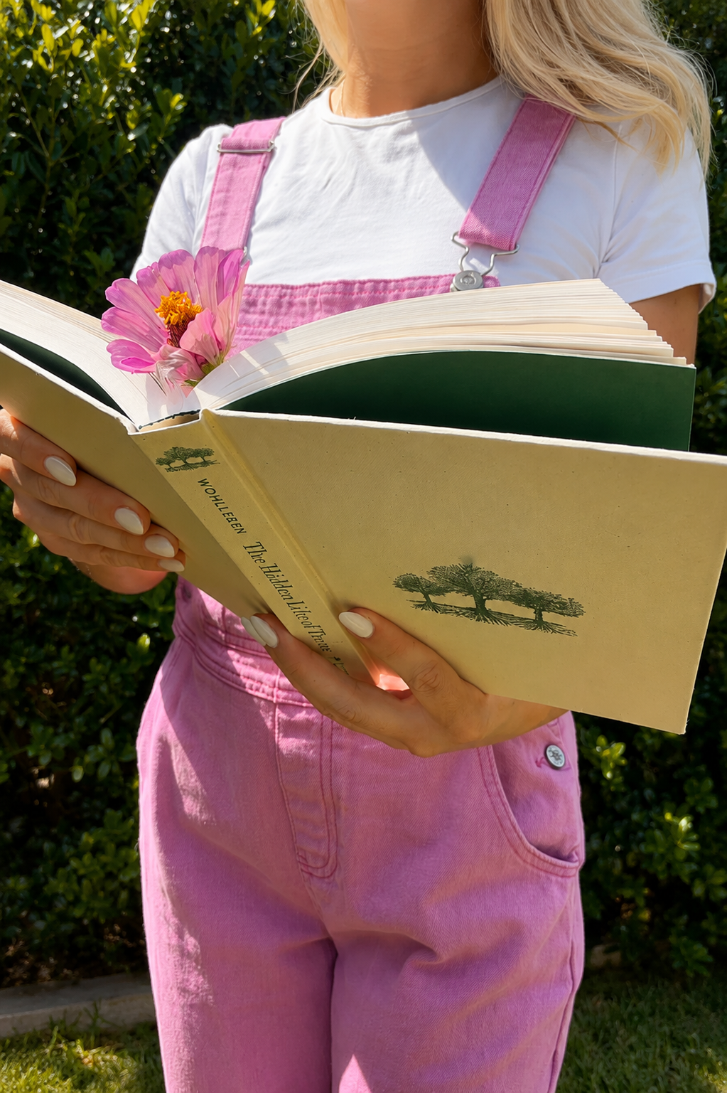 A woman wearing pink overalls and a white t-shirt holds a book, with a pink flower tucked into the open pages, standing outdoors with green foliage in the background.