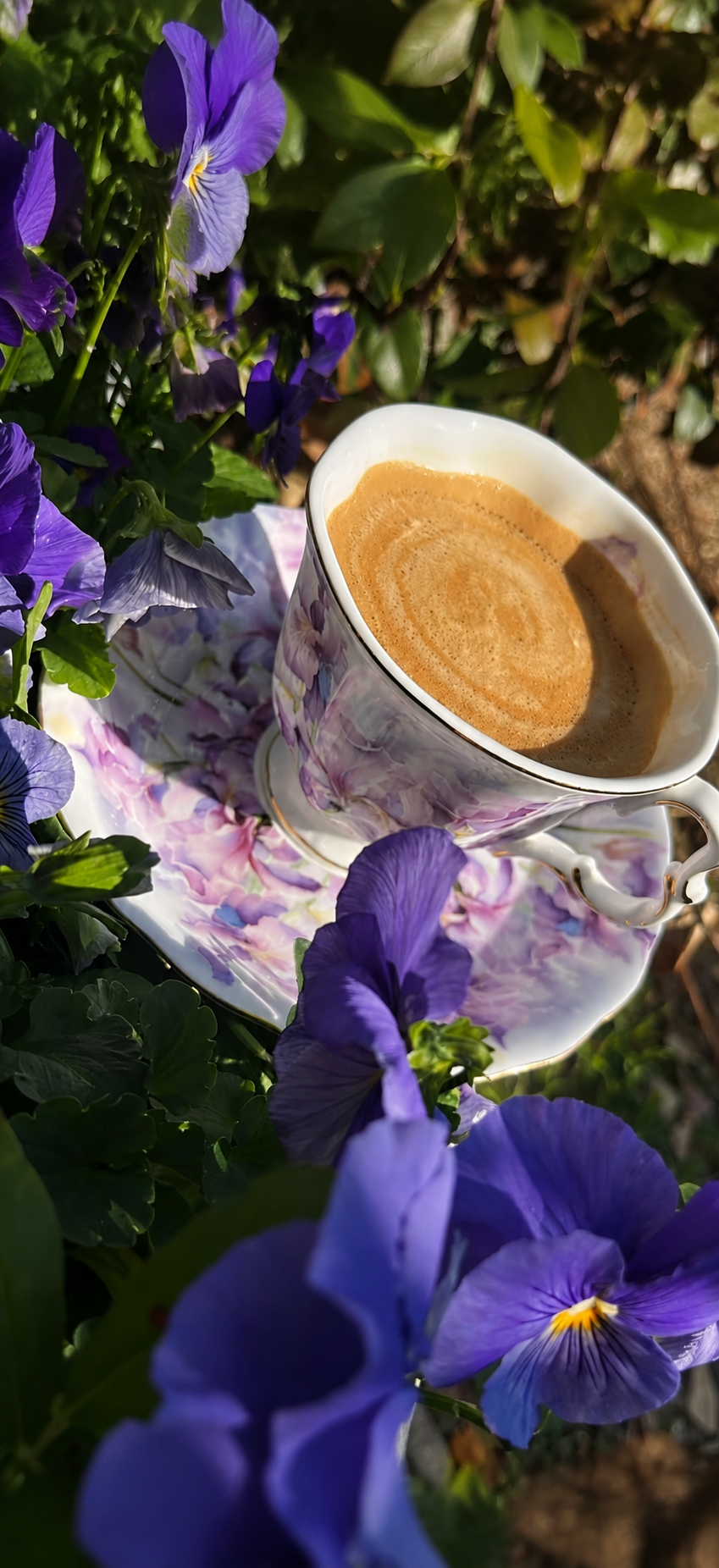 A marble-patterned teacup with a floral design, filled with coffee, placed on a matching saucer among purple pansy flowers with green leaves, outdoors in sunlight.