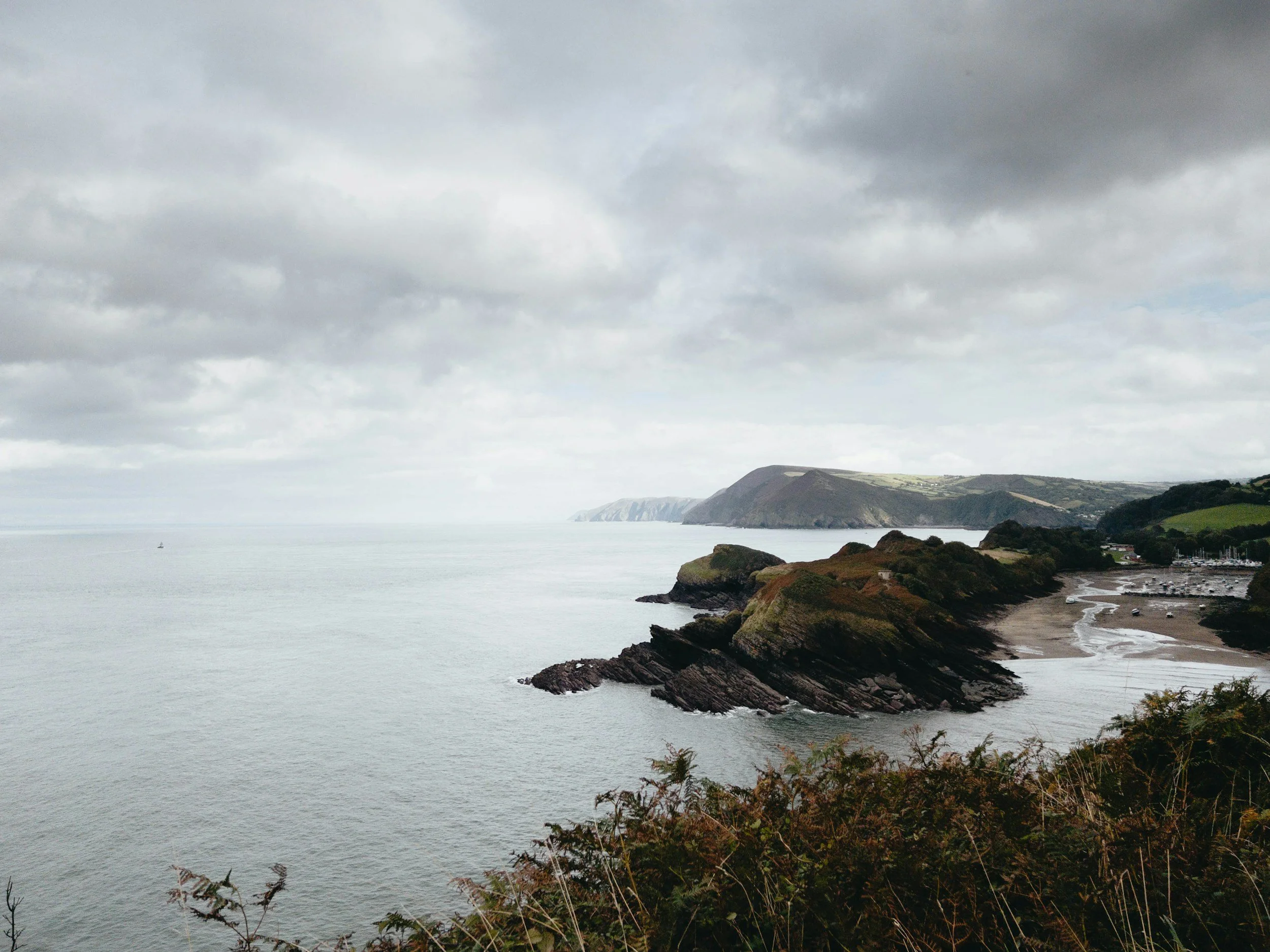 Coastal landscape with cloudy sky, hills, and rocky shoreline by the ocean, with some vegetation in the foreground.