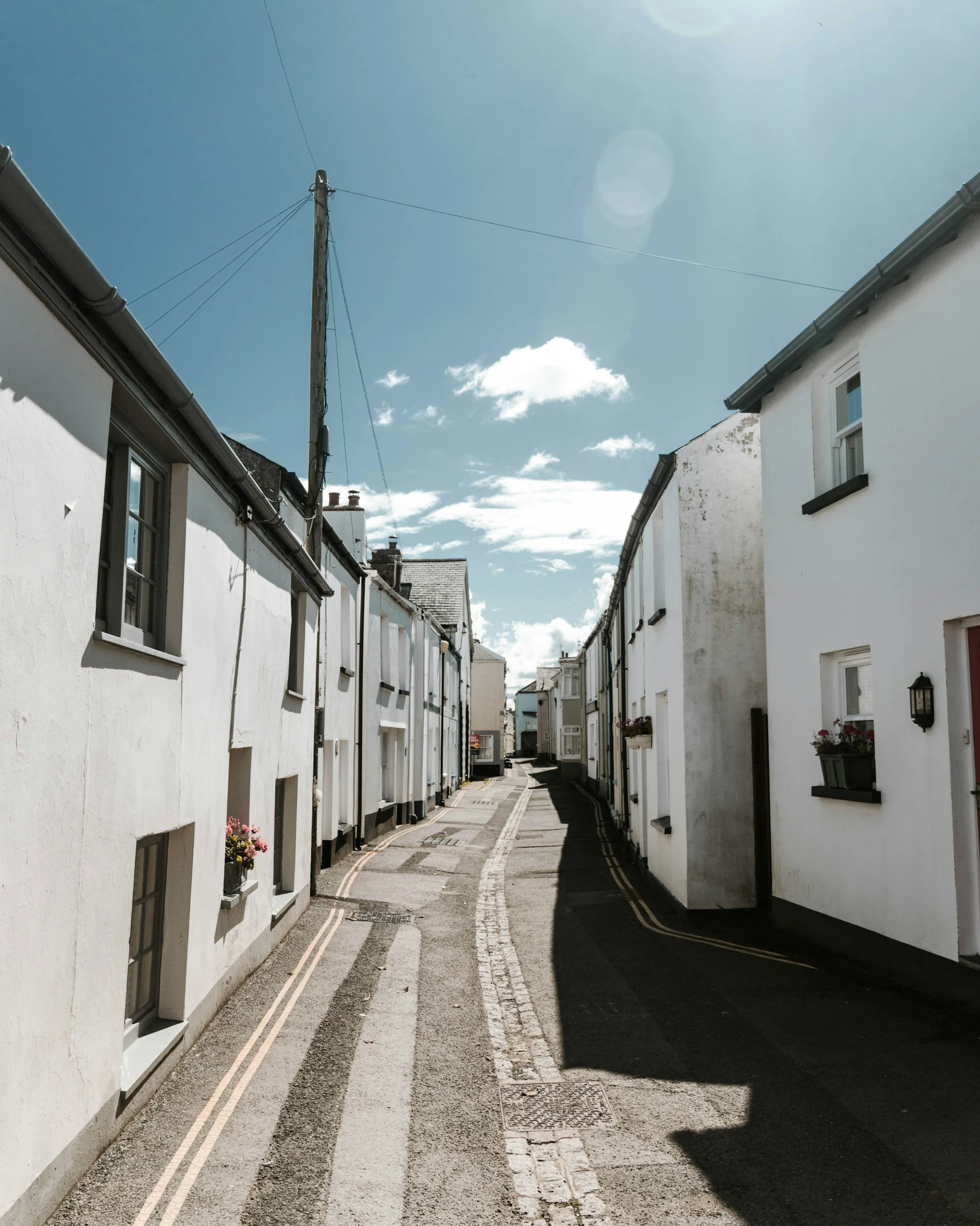 A narrow street between white residential buildings with dark roofs, a large utility pole with wires extends into a bright, mostly clear sky with a few clouds.