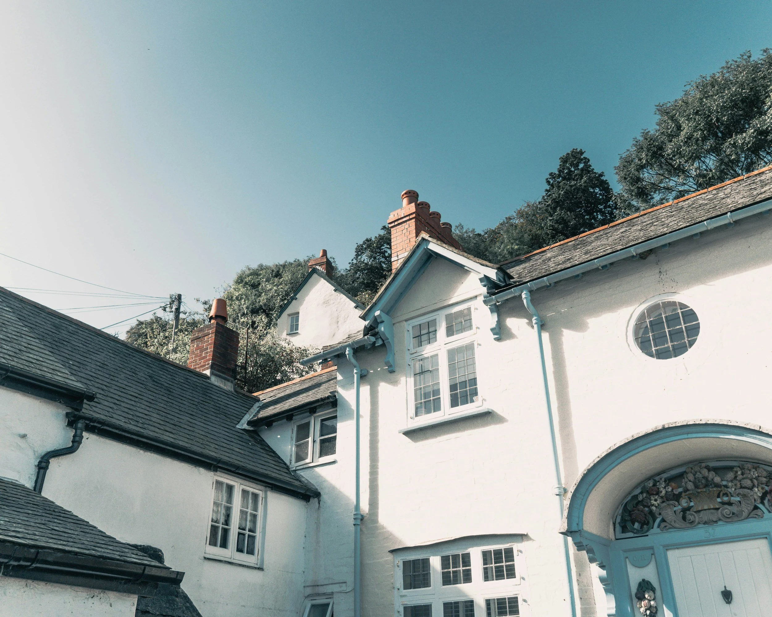 A charming white house with a steep slate roof and multiple chimneys set against a bright blue sky and trees.