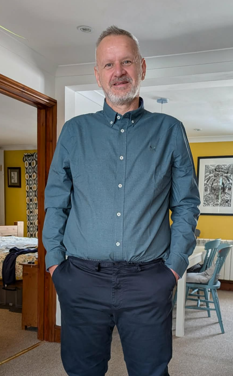 A smiling middle-aged man with a beard, standing indoors in front of a yellow and white room with furniture. He is wearing a teal button-up shirt and dark pants.