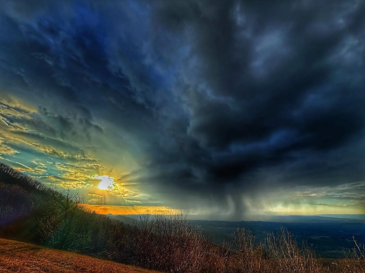 A dramatic stormy sunset over a hilly landscape with dark clouds, sunlight breaking through, and rain falling in the distance.