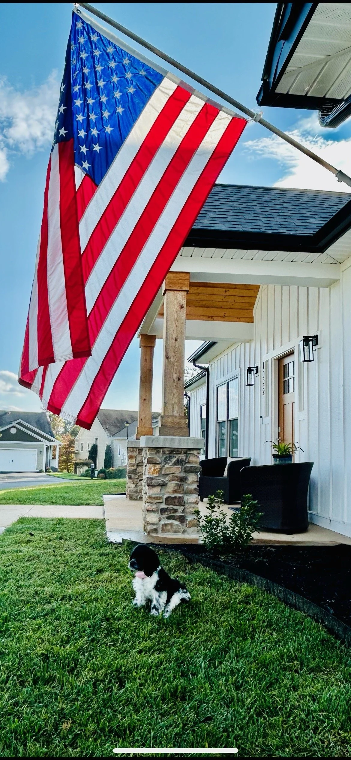 An American flag hanging outside a modern house with patio furniture and a small black and white puppy sitting on the grass.