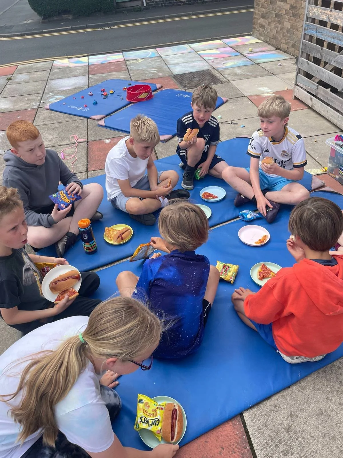 Children sitting on a blue mat outside, having a picnic with hot dogs, pizza, and snacks, on a paved area near a street and brick wall.