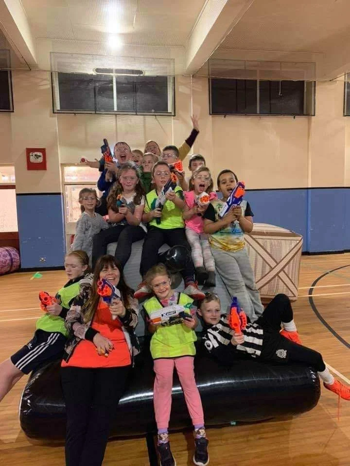 Group of children and teenagers holding toy guns, posing on an inflatable obstacle in a gymnasium or indoor sports hall, with some smiling and making playful poses.