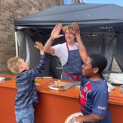 Three children smiling and giving high fives at an outdoor food stand. One child is behind a counter with a black canopy, serving food in a tray.