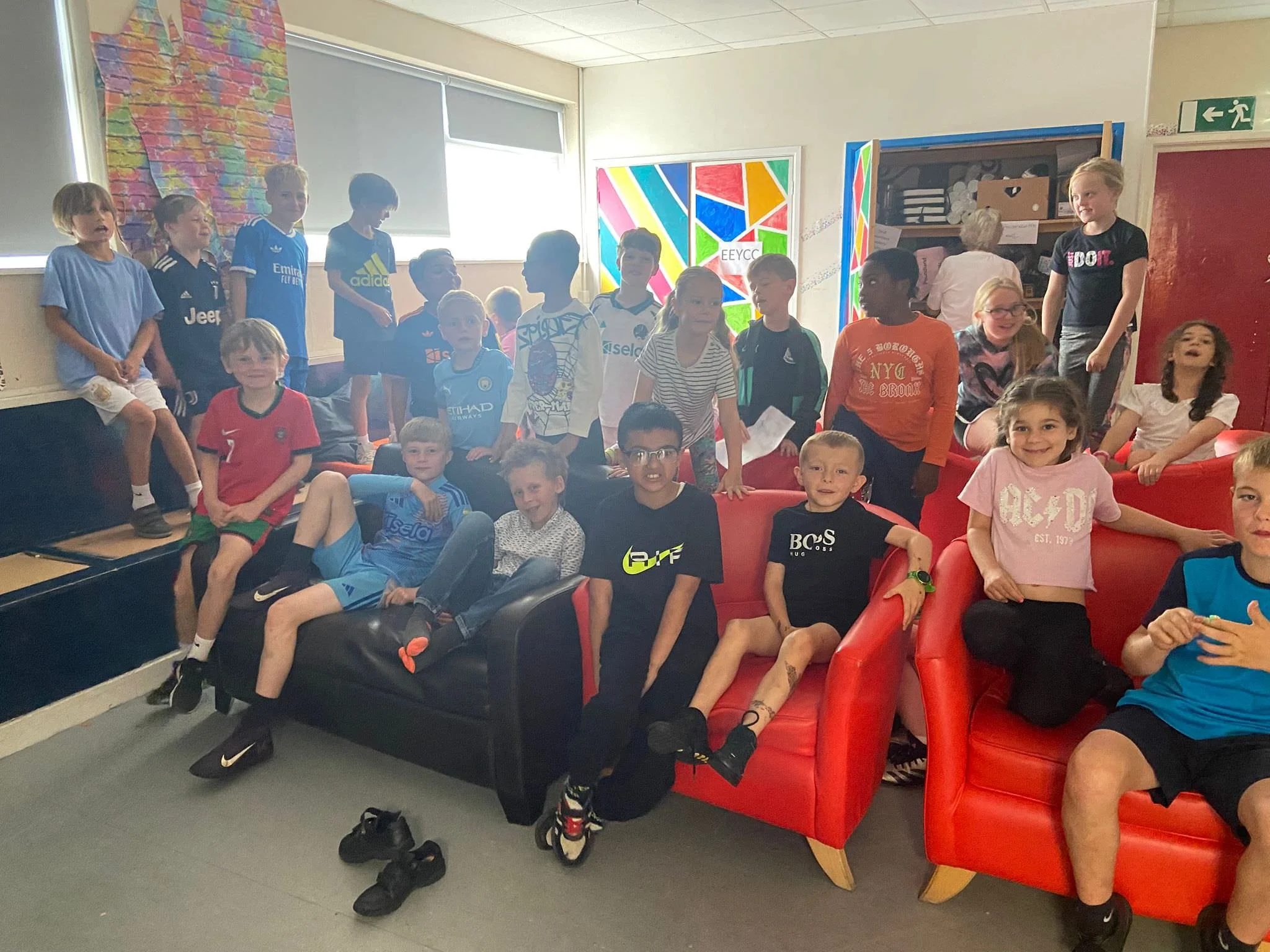 A group of children sitting and standing in a room with colorful wall art and red seating, some smiling, with shoes on the floor.