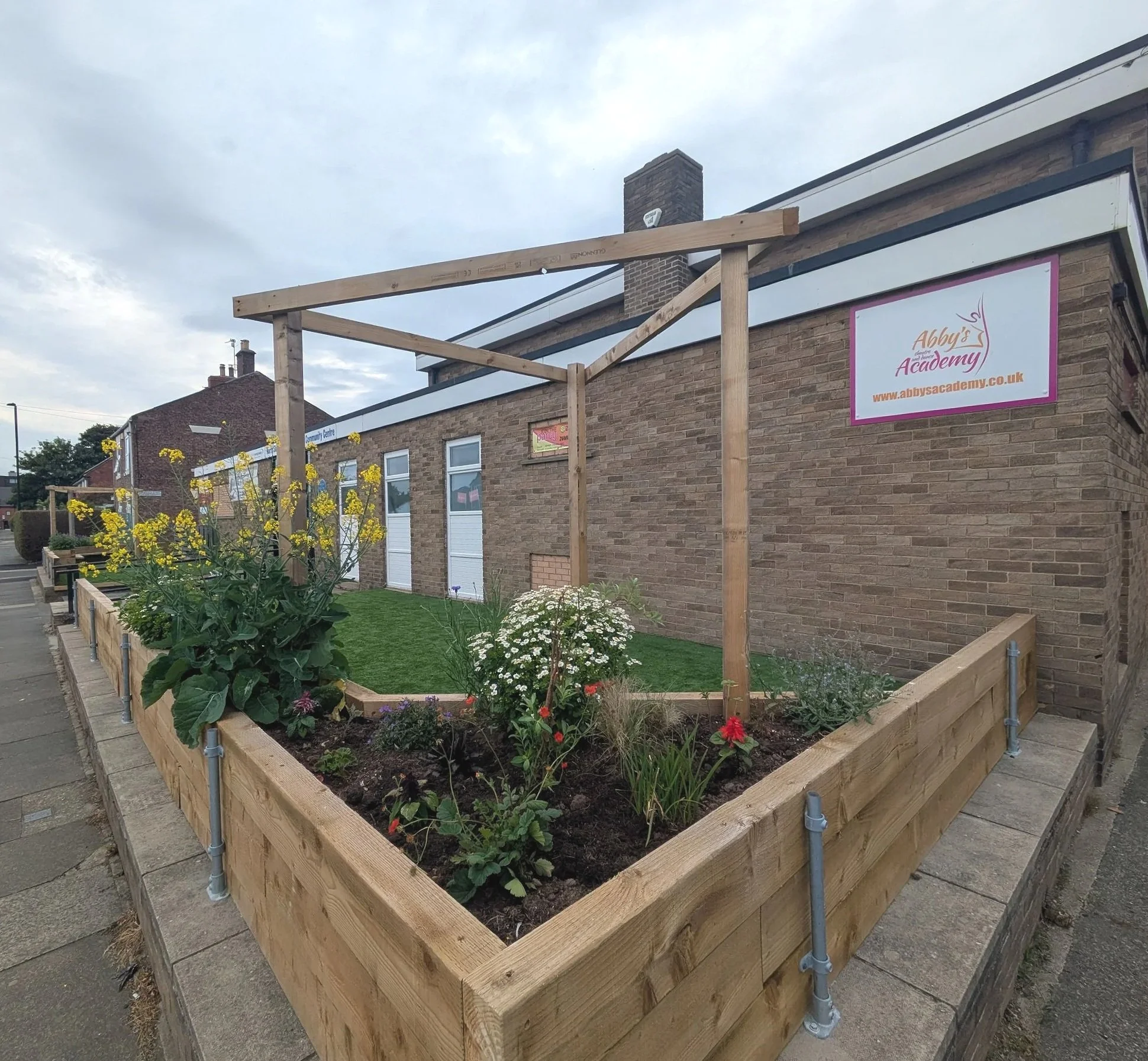 A raised garden bed made of wooden planks filled with flowers and plants, placed on the sidewalk outside a brick building under construction.