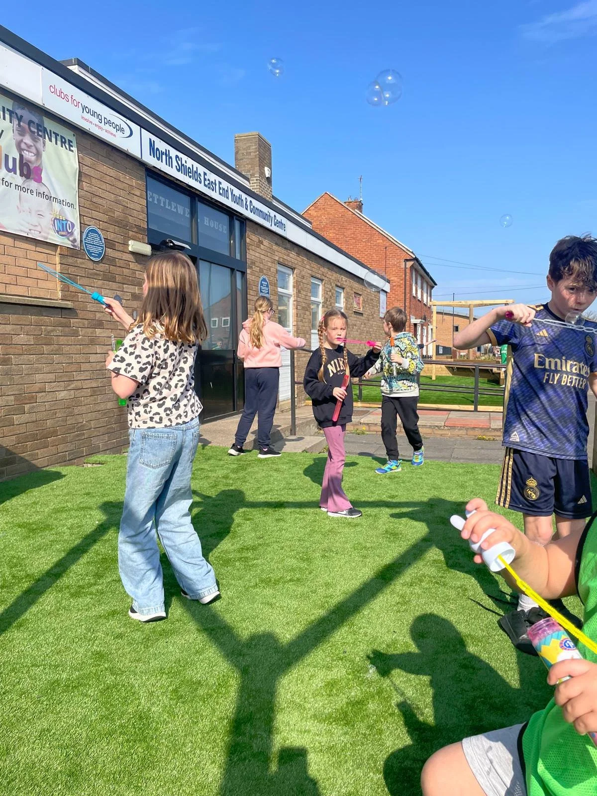 Children playing with bubble wands outside a community center on a sunny day.