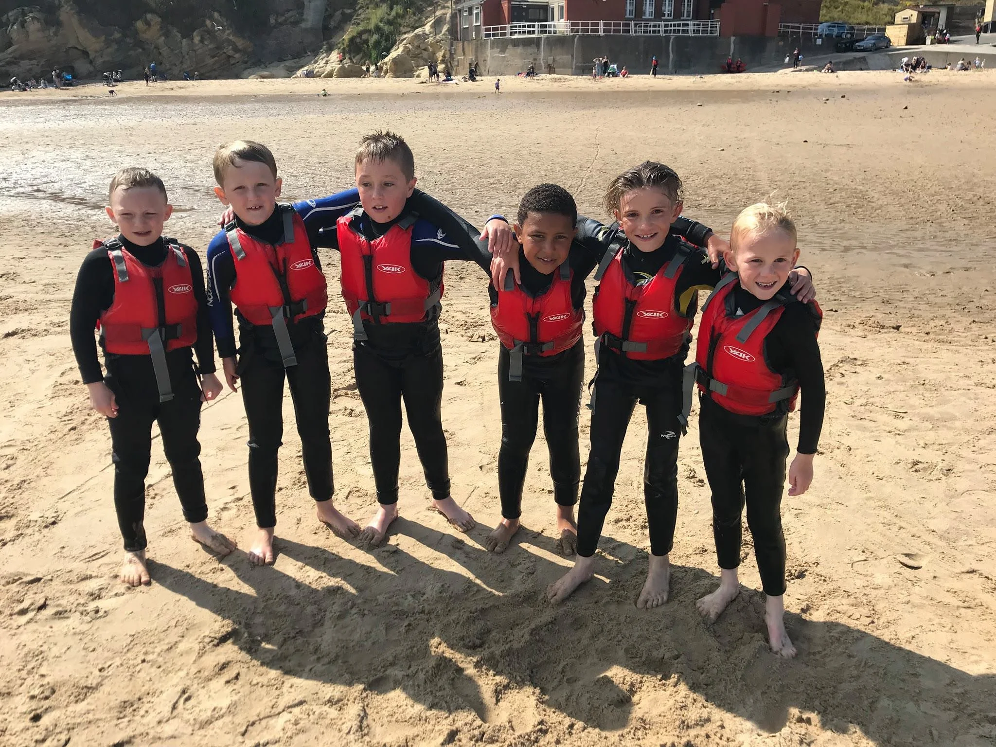Group of six children standing barefoot on the sandy beach, wearing black wetsuits and red life jackets, smiling for the camera.