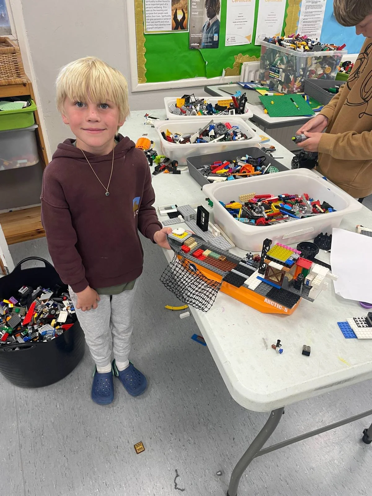 A young boy with blonde hair wearing a brown hoodie, light gray pants, and blue Crocs standing at a table filled with LEGO pieces. He is holding a partially assembled LEGO model. Behind him are other LEGO pieces and another child working on a different LEGO project in a classroom setting with posters on the wall.