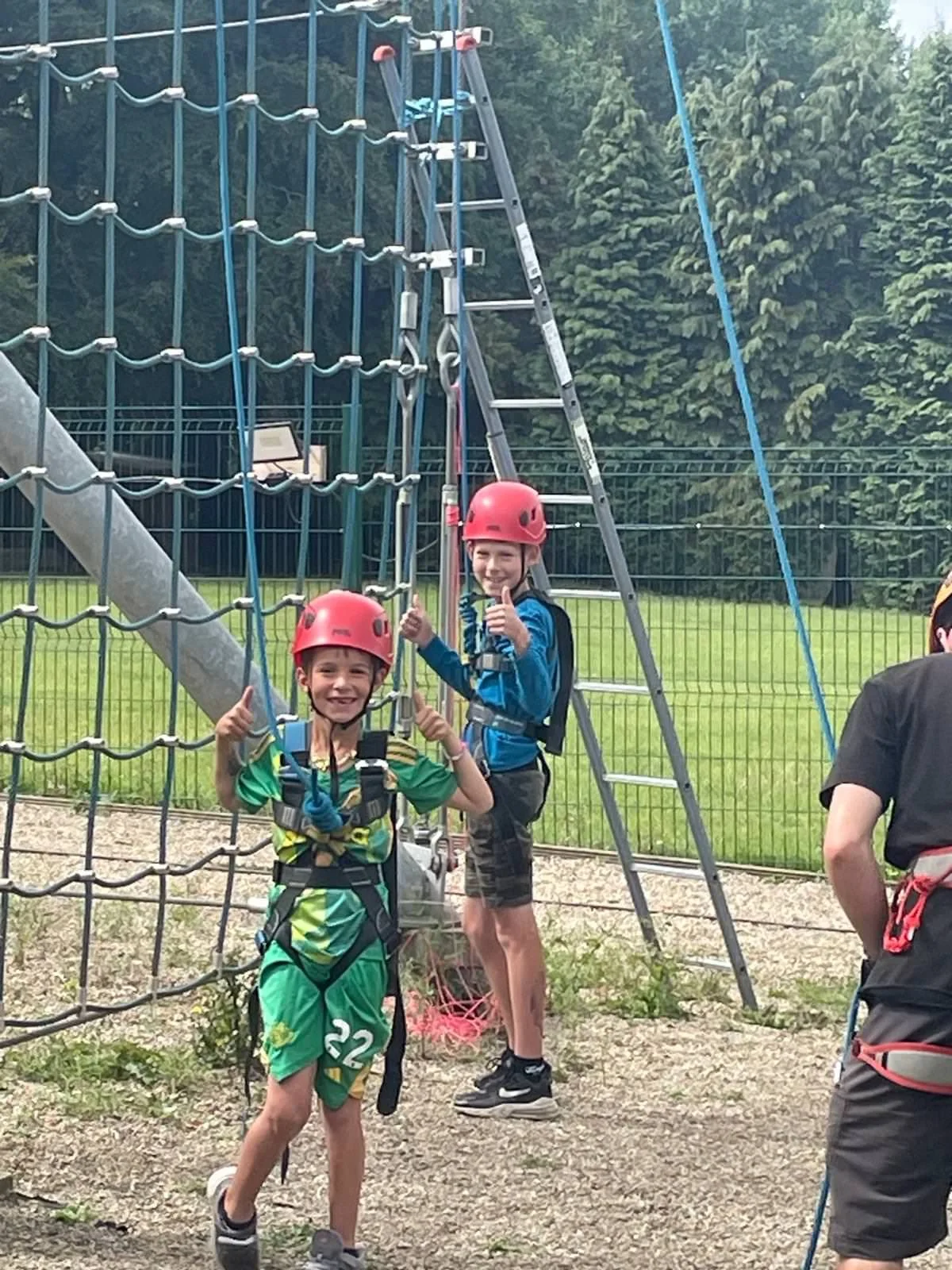 Two boys wearing helmets and harnesses at an outdoor ropes course, smiling and giving thumbs-up, with safety equipment and ladder in the background.