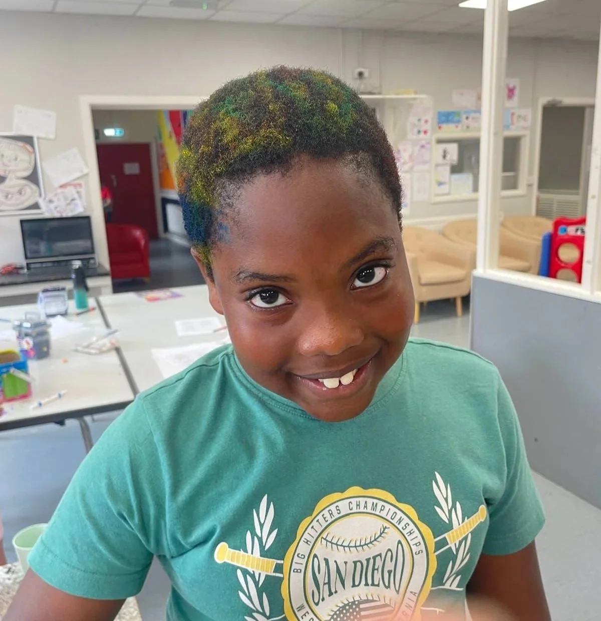 A young girl with short, curly hair painted with multiple bright colors, smiling in an indoor recreational or classroom setting.