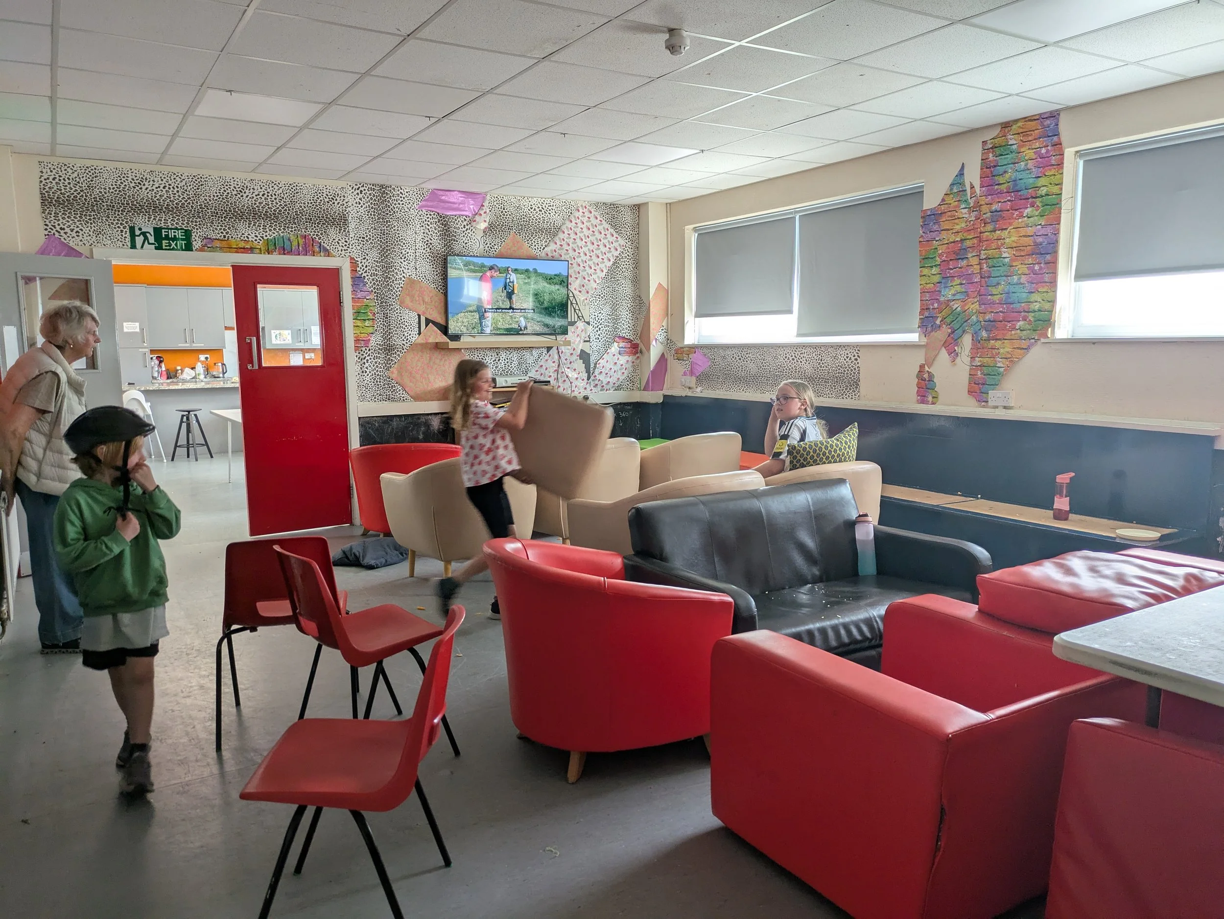 A room with colorful wallpaper on part of the walls, featuring a map design and a leopard print pattern, with a television on the wall displaying a scene of two people walking outdoors. There are four children and an adult, some standing and some sitting, in the foreground. The room has windows with roller blinds and red and black chairs, as well as beige sofas. The background includes a kitchen area with gray cabinets and a door with a fire exit sign above it.