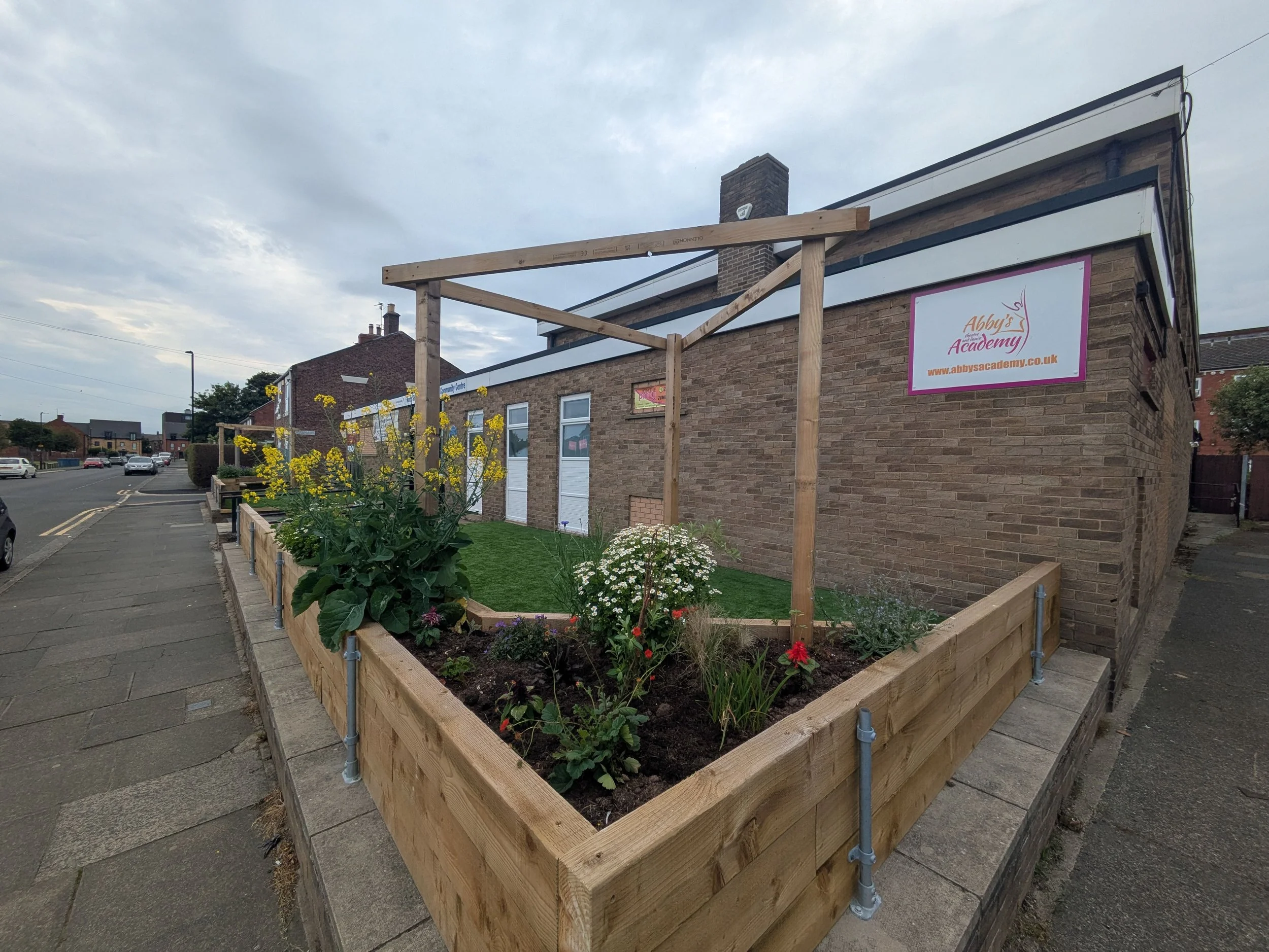 A raised wooden garden bed with various flowers and plants, situated on a sidewalk in front of a brick building with signs, with a cloudy sky overhead.