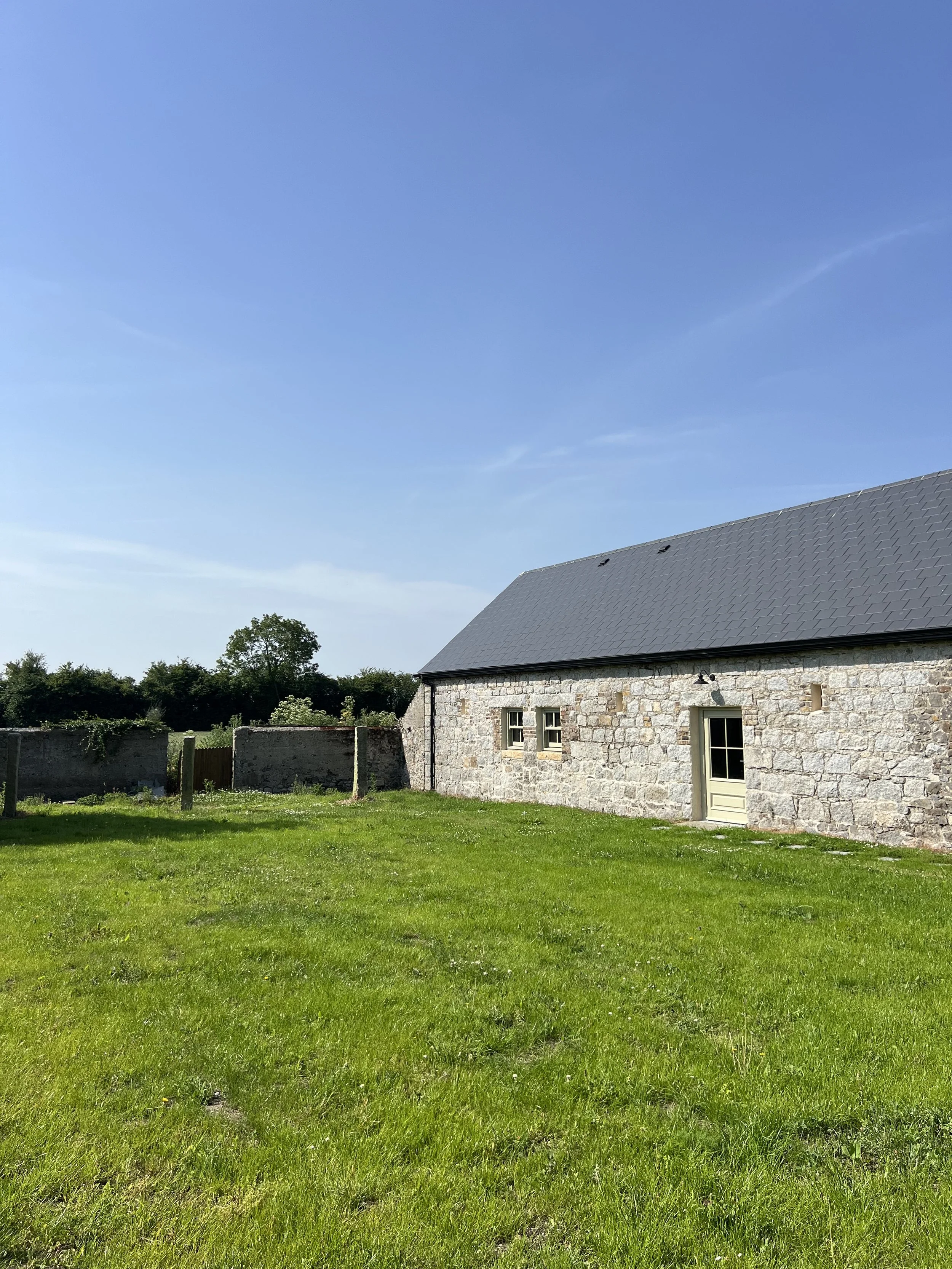 A stone house with a gray shingled roof, a green door, and three windows, set in a grassy yard with a clear blue sky overhead and trees in the background.