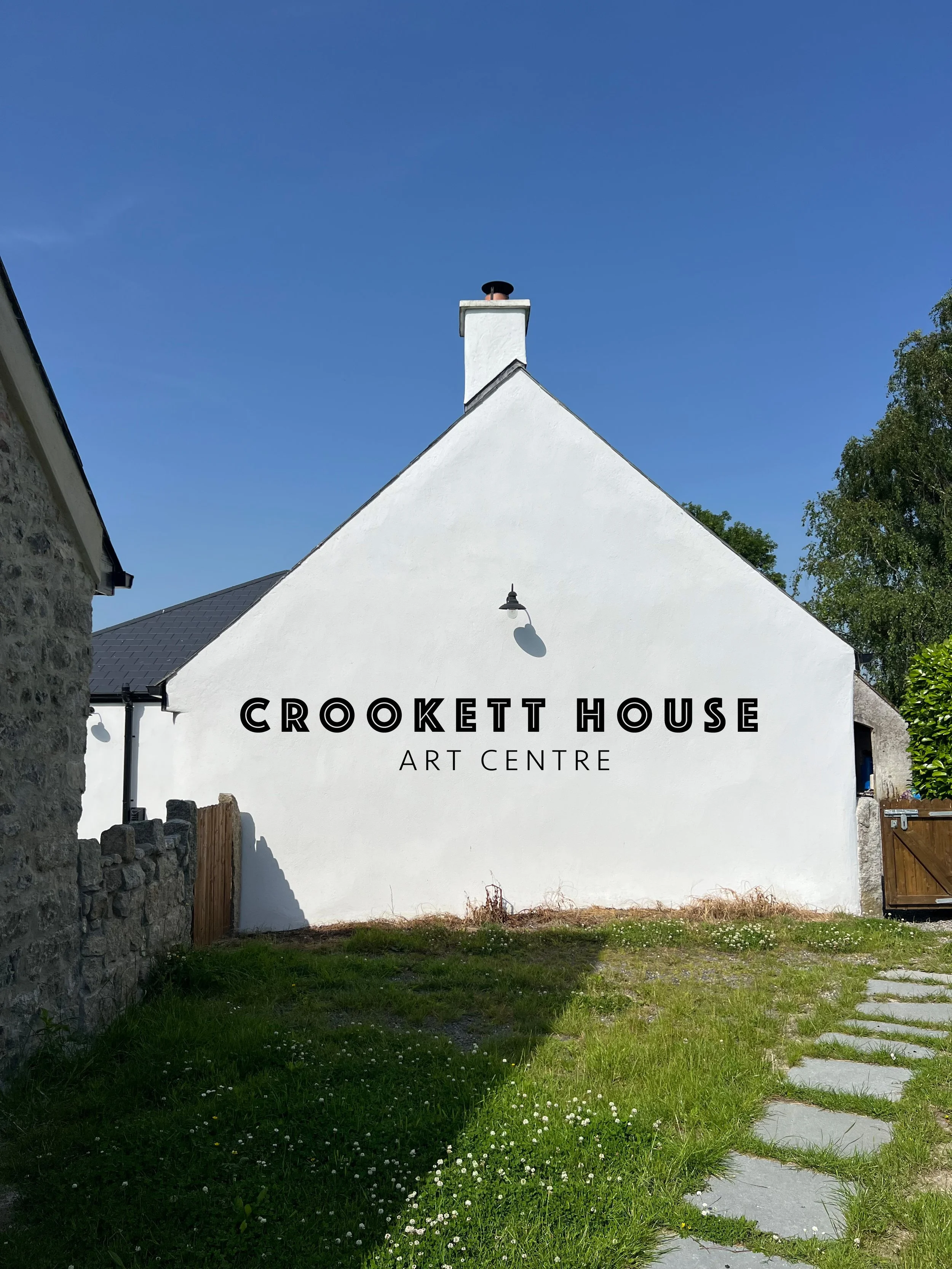 White building with a triangular roof and chimney, sign reading 'Crookett House Art Centre,' stone wall on the left, grassy area with stepping stones on the right, blue sky, and trees in the background.