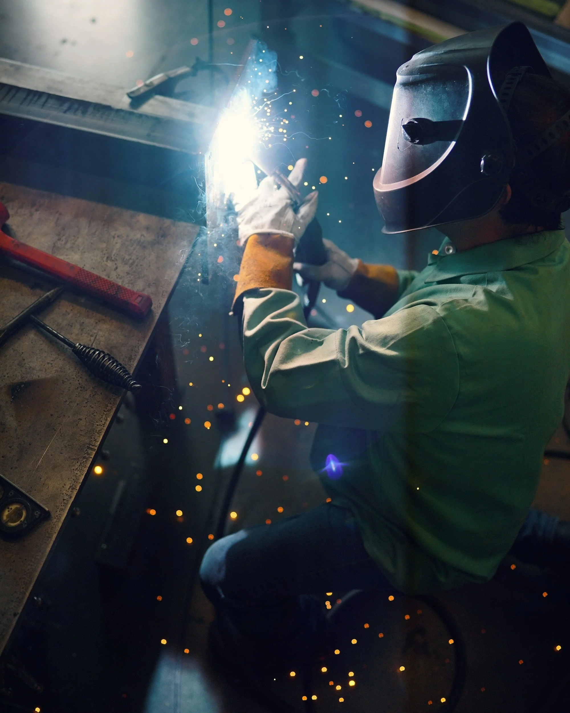 A person welding metal, wearing a protective helmet and gloves, with sparks flying around from the welding process.