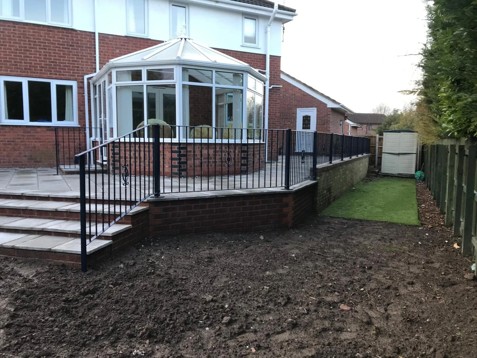 Backyard with a raised patio, brick steps, black metal railing, and a conservatory attached to a brick house. Some areas of the yard are bare dirt, and others are covered in green grass. A garden shed and trees are visible in the background.
