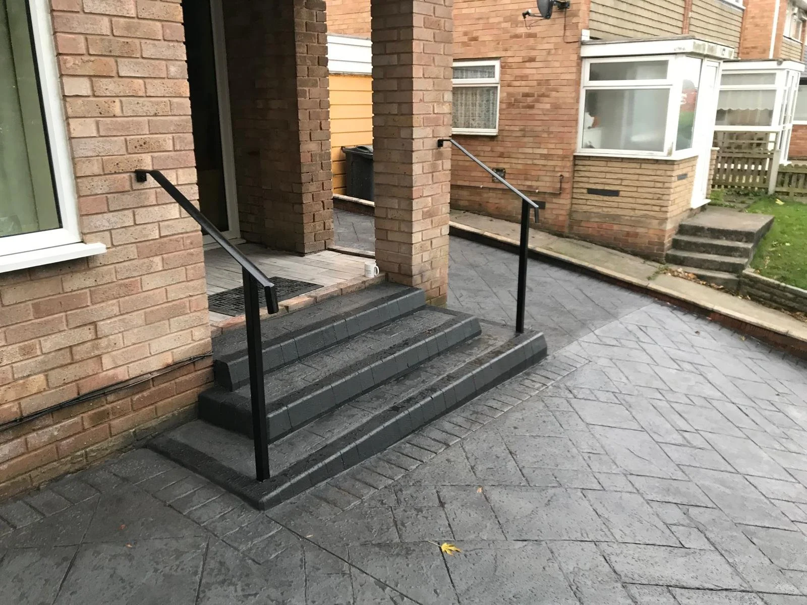 An entrance with a small staircase featuring dark gray bricks, black handrails, and rubber doormat mats. The building is made of red brick with a partial yellow siding wall. There are steps leading to a side door and another set of steps on the right
