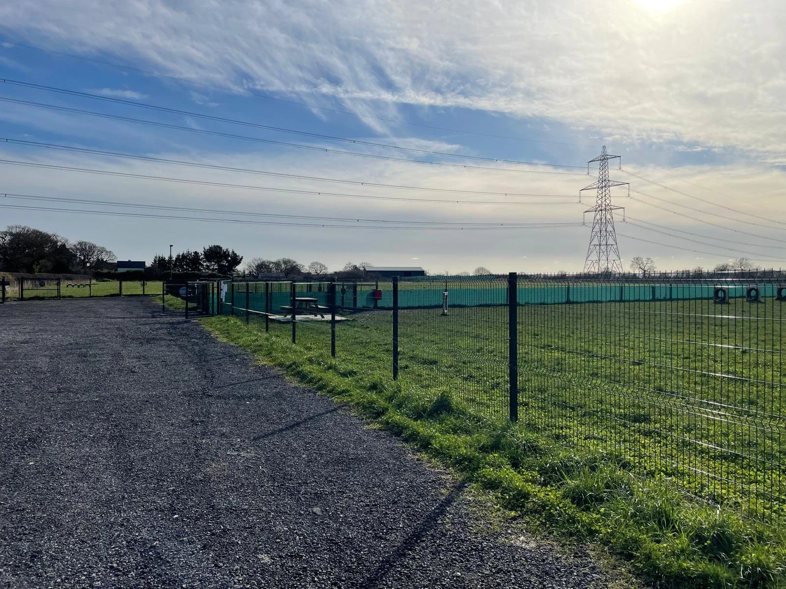 A grass field enclosed by a black metal fence with a gravel path running alongside it. There are some picnic tables, a small shed, and large power lines with a transmission tower in the background under a partly cloudy sky.