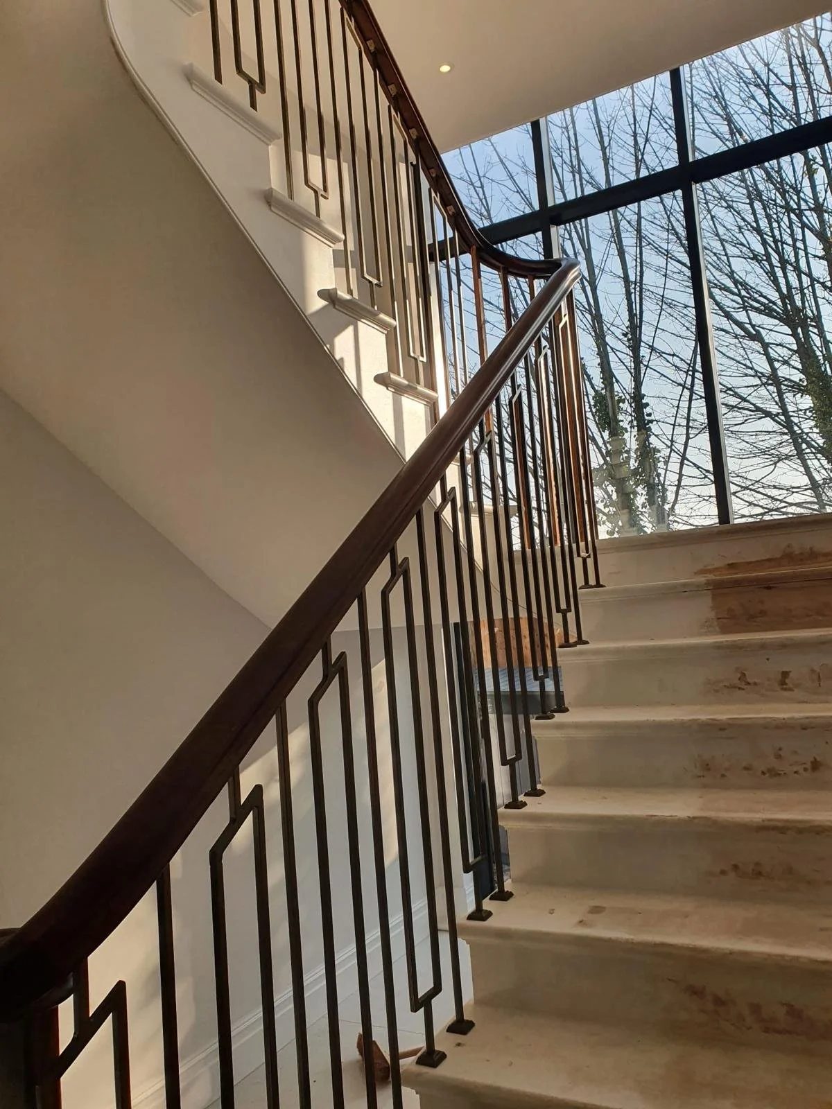 Indoor view of a staircase with wooden and metal railing, marble steps, large glass window with view of trees outside, and sunlight streaming in.