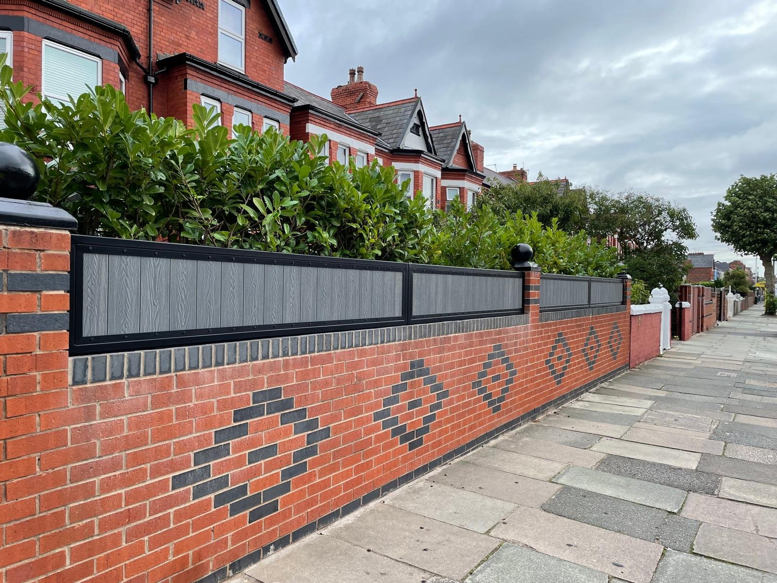 A brick wall with decorative diamond-shaped patterns, topped with black and gray fencing, along a sidewalk in front of red brick residential buildings and green bushes.