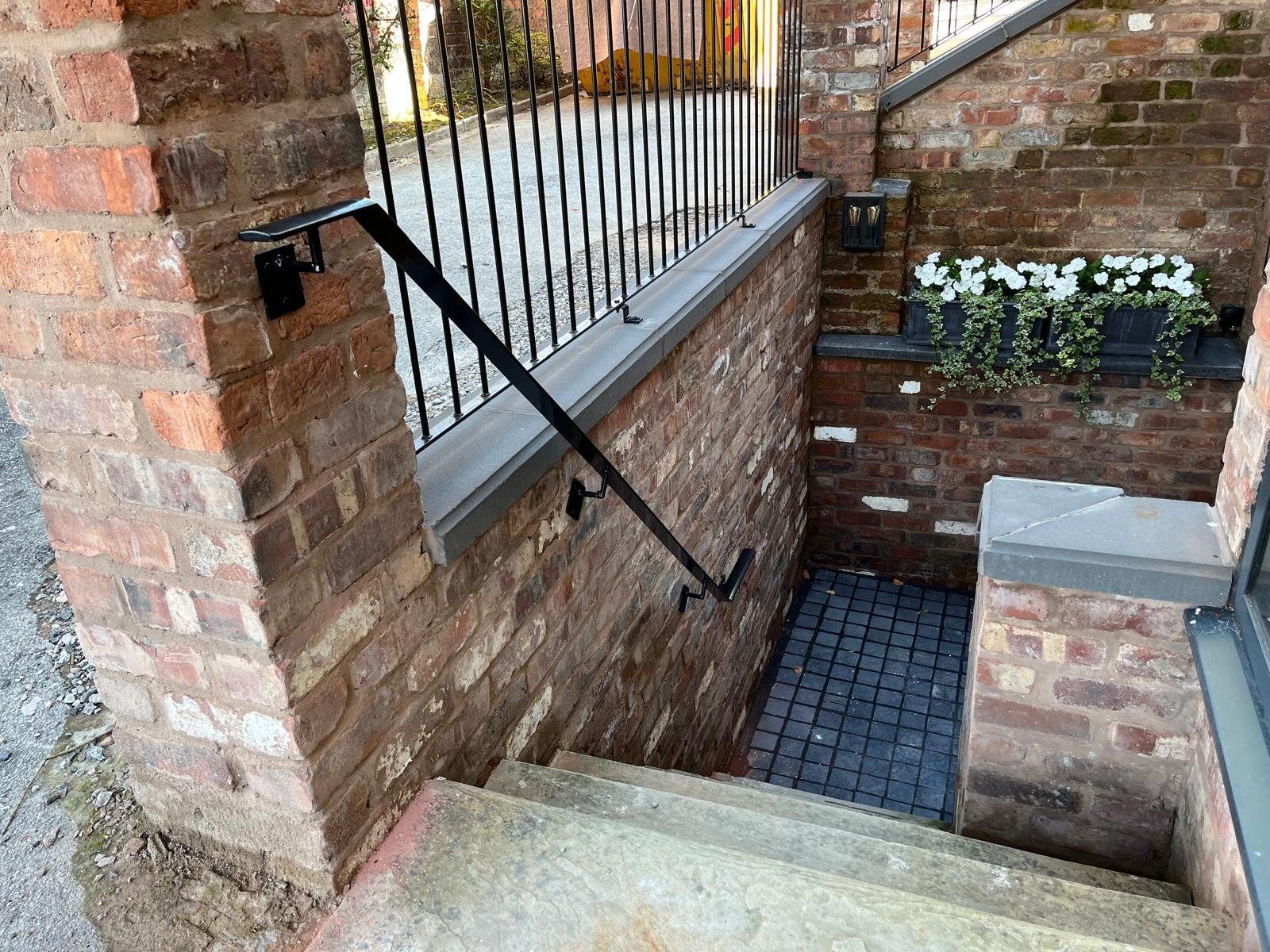 Brick staircase leading down to a basement entrance with a black metal handrail on the left side, a brick wall with a flower box filled with white flowers on the right side, and an outdoor lantern attached to the brick wall.