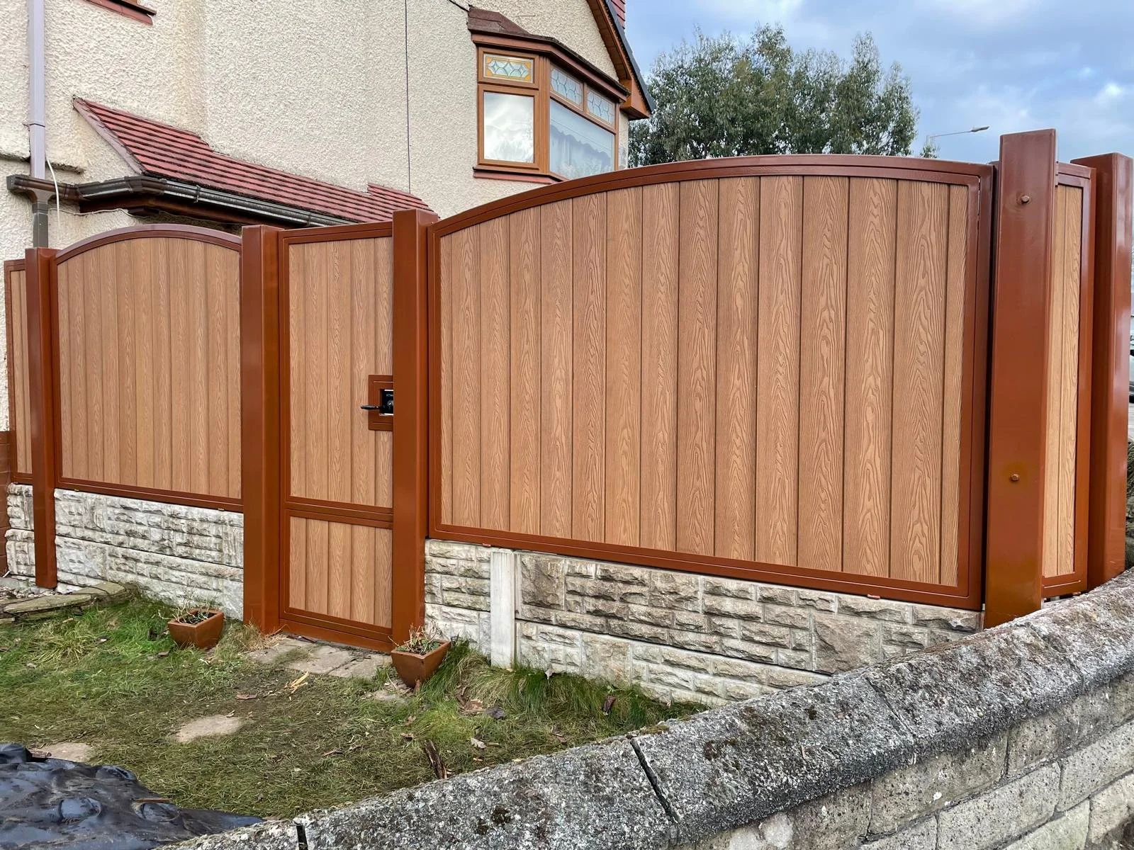 Wooden privacy fence with a matching gate in front of a house with a stone foundation and a tree in the background.