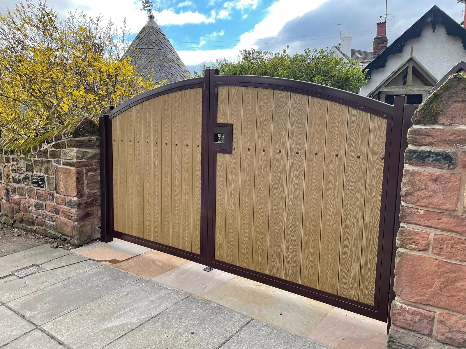 A wooden gate with a curved top, set between stone pillars, with a small square locking mechanism in the middle.